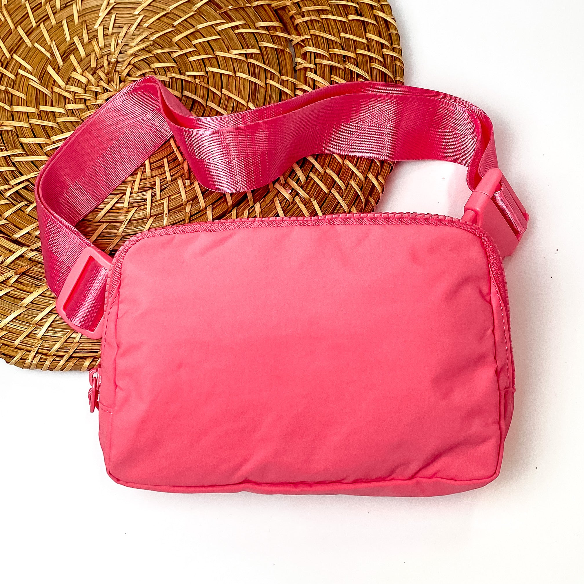 Pictured is a rectangle fanny pack with a top zipper with tassel in watermelon pink. This bag also includes a watermelon pink strap and watermelon pink accents. This bag is pictured on a white and brown patterned background. 
