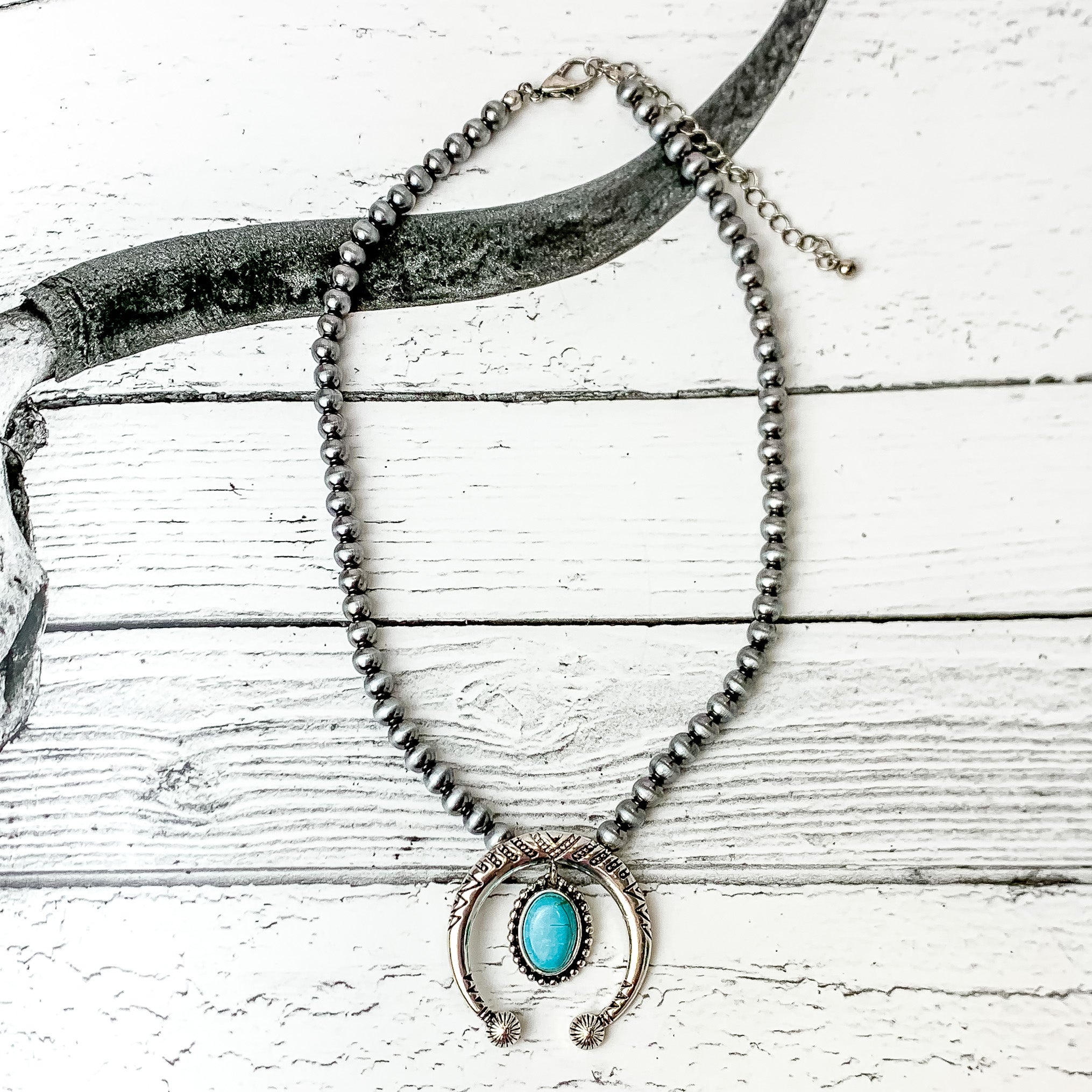Squash blossom in silver faux navajo pearls with a dangling oval turquoise stone. Pictured on a black and white western photo in the background.