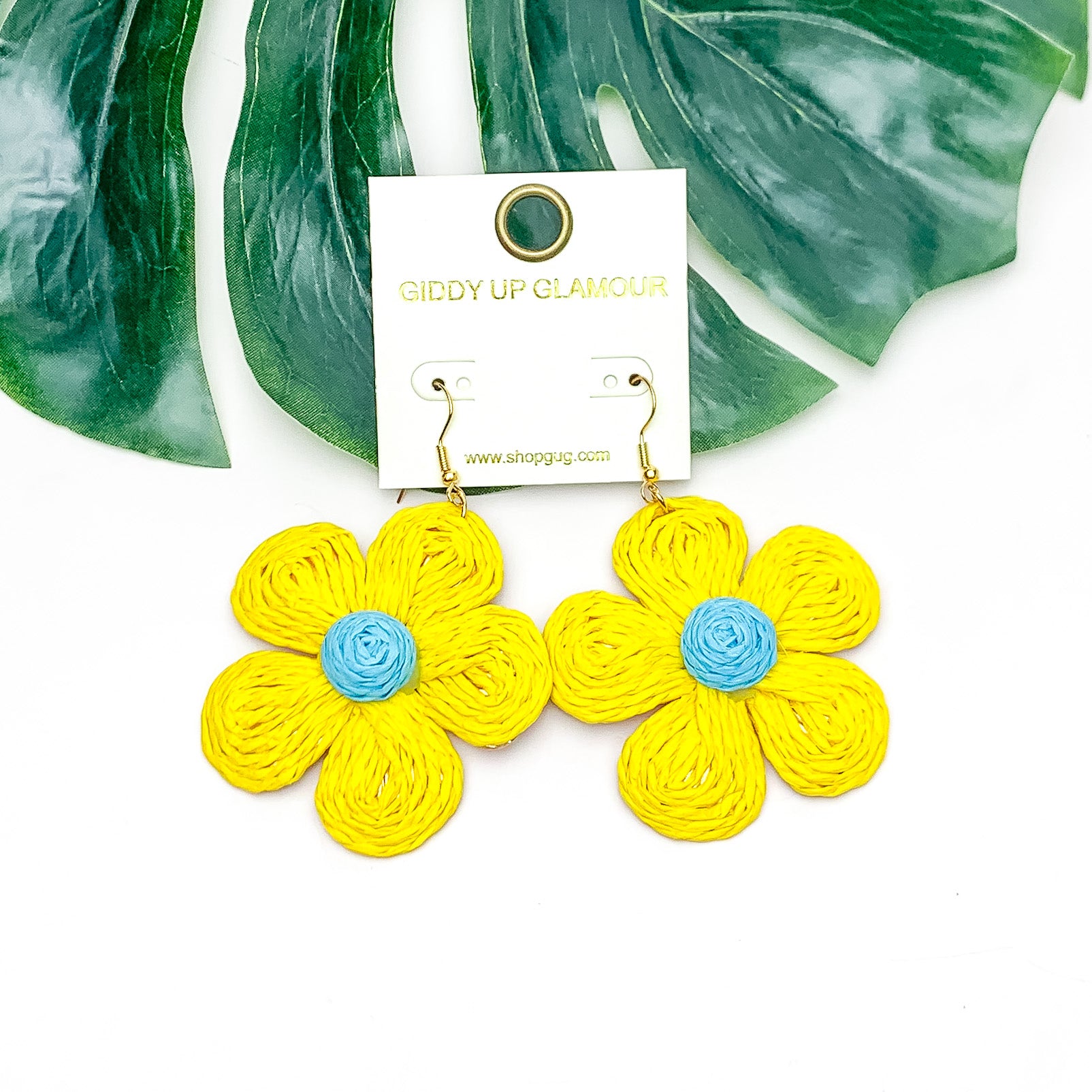 Darling Daisy Raffia Wrapped Flower Earrings in Yellow. Pictured on a white background with the earrings laying on a large leaf.
