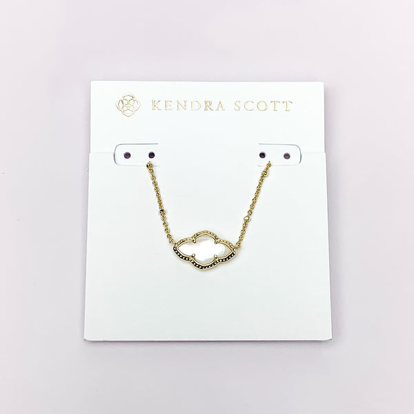 Gold chain necklace with a gold quatrefoil pendant and ivory mother of pearl. This necklace is pictured on a white necklace holder on a white background. 