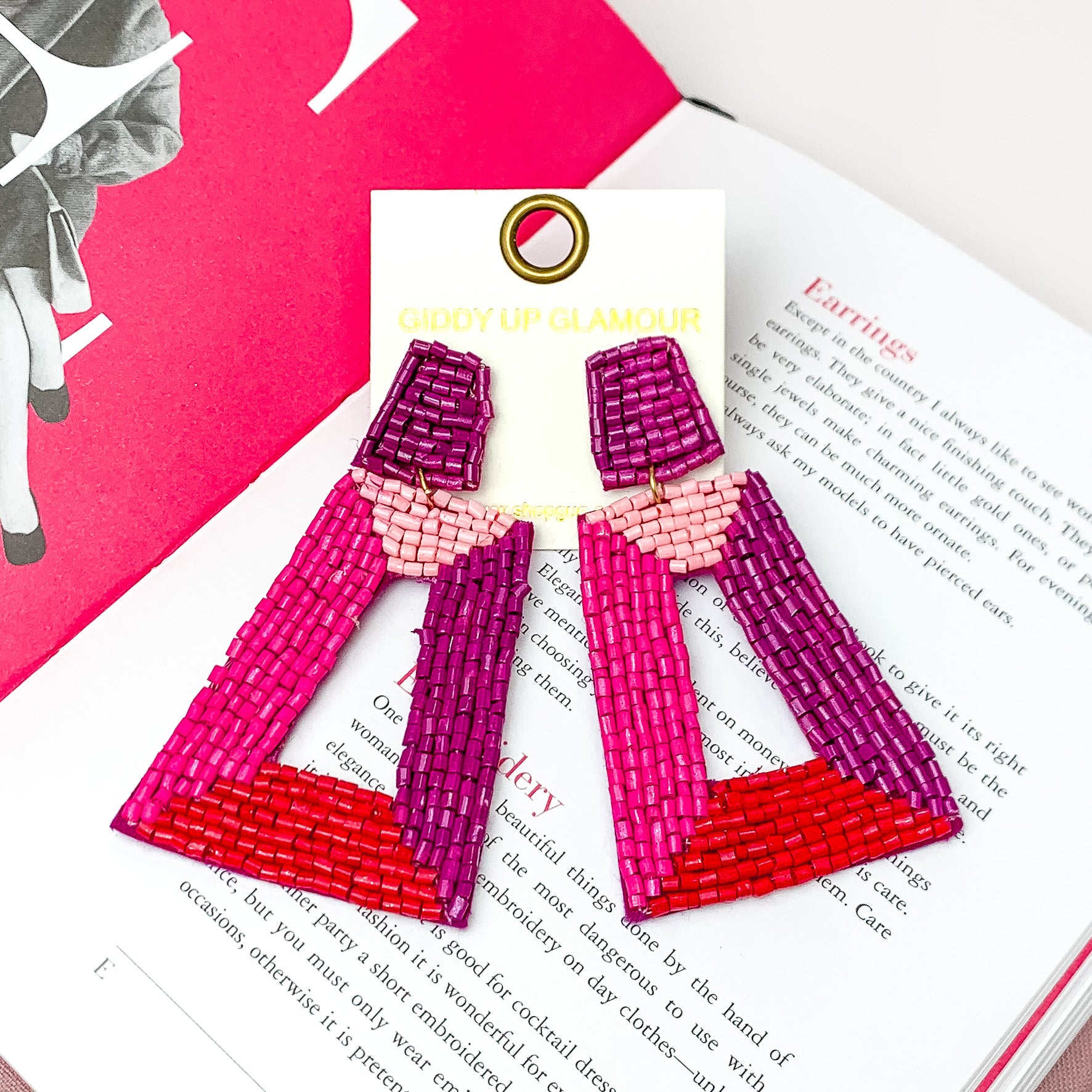Beaded Rectangle Designed Earrings in Pink, Red, and Purple. Pictured on a white background with a book behind the earrings. 