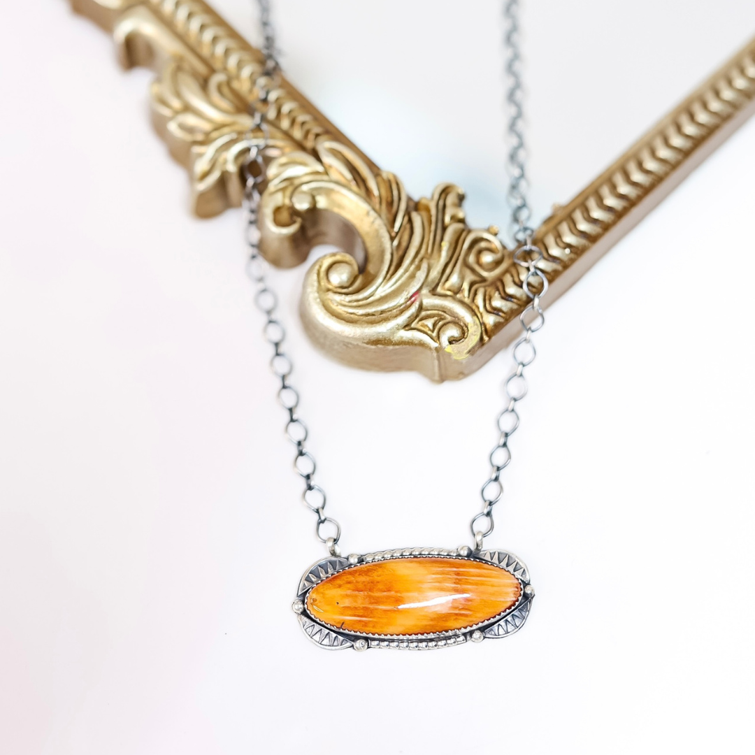 Sterling silver necklace with a long oval orange spiny oyster pendant, handmade by Navajo artist Tia Long, styled on a white background with a gold mirror.