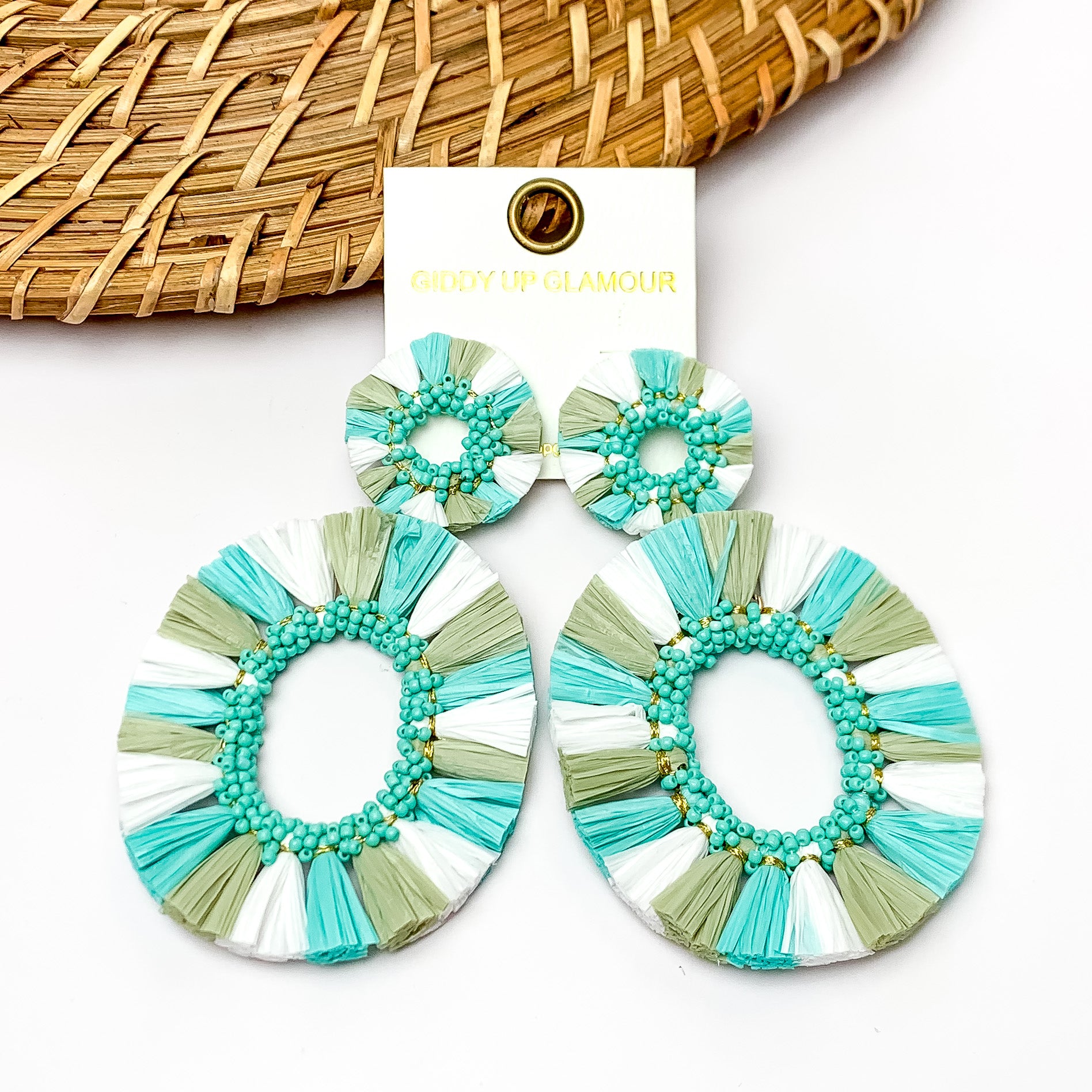 Siesta Keys Raffia Wrapped Open Oval Earrings in Turquoise, Green, and White. Pictured on a white background with wood like decoration in the top left corner.