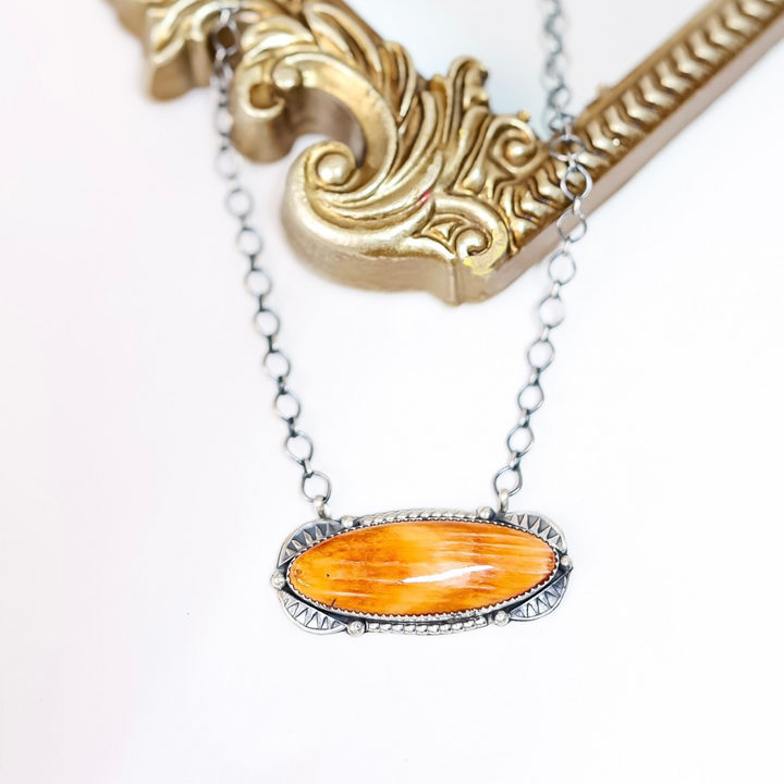 Sterling silver necklace with a long oval orange spiny oyster pendant, handmade by Navajo artist Tia Long, styled on a white background with a gold mirror.