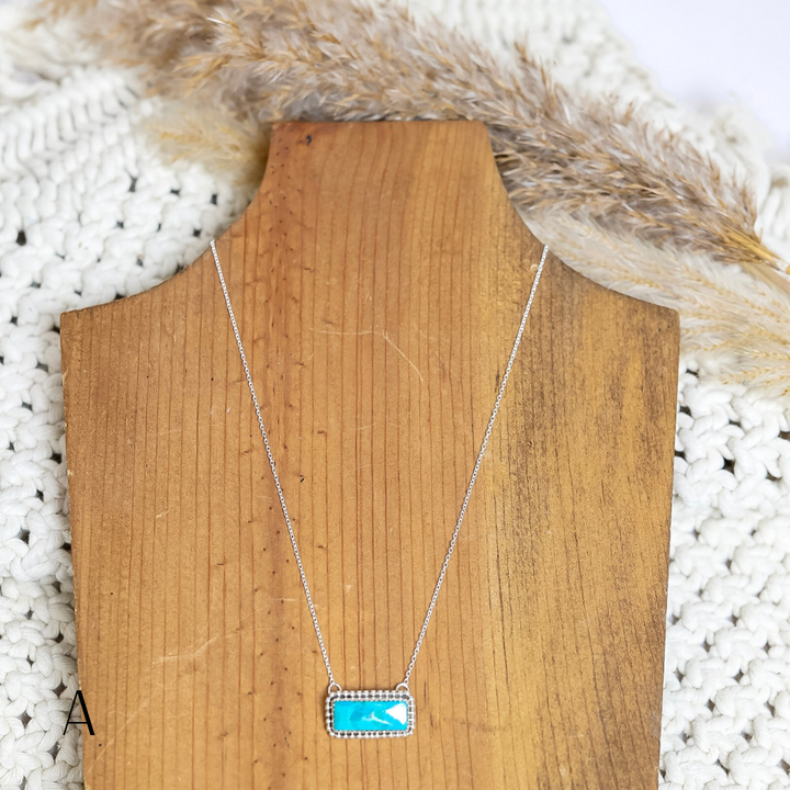 Handmade Navajo necklace displayed on a wooden stand, featuring a rectangular turquoise stone set in a detailed silver bezel on a delicate silver chain, with neutral textured background and dried pampas grass for styling.
