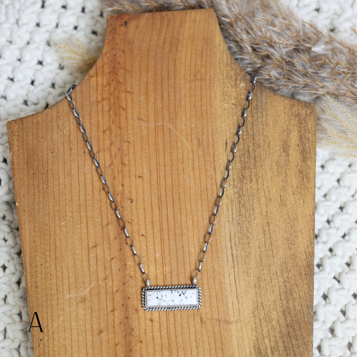 Handmade Navajo necklace displayed on a wooden stand, featuring a rectangular white buffalo stone set in a detailed silver bezel on a delicate silver chain, with neutral textured background and dried pampas grass for styling.