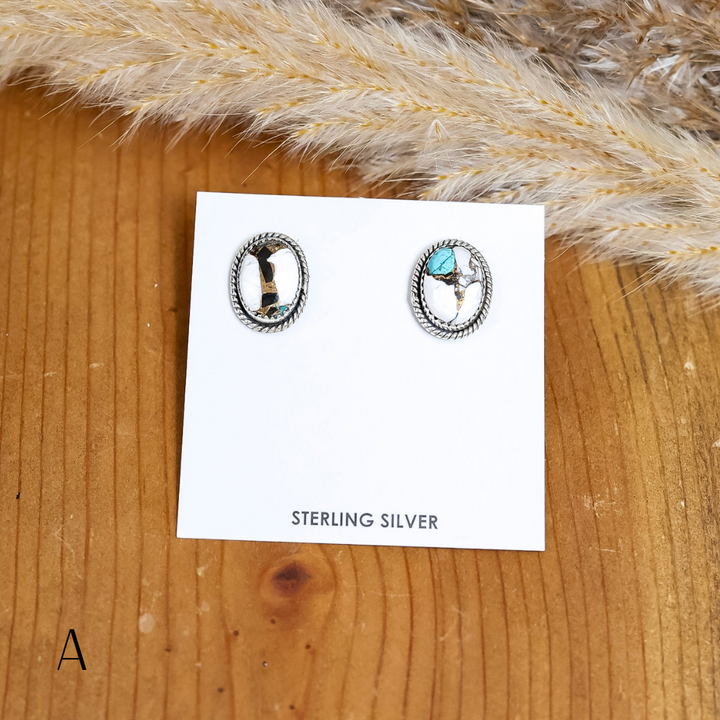 Pair of oval Navajo turquoise stud earrings with silver rope-edge detail, displayed on a white card labeled “Sterling Silver,” set against a wooden background with pampas grass and a knit texture.