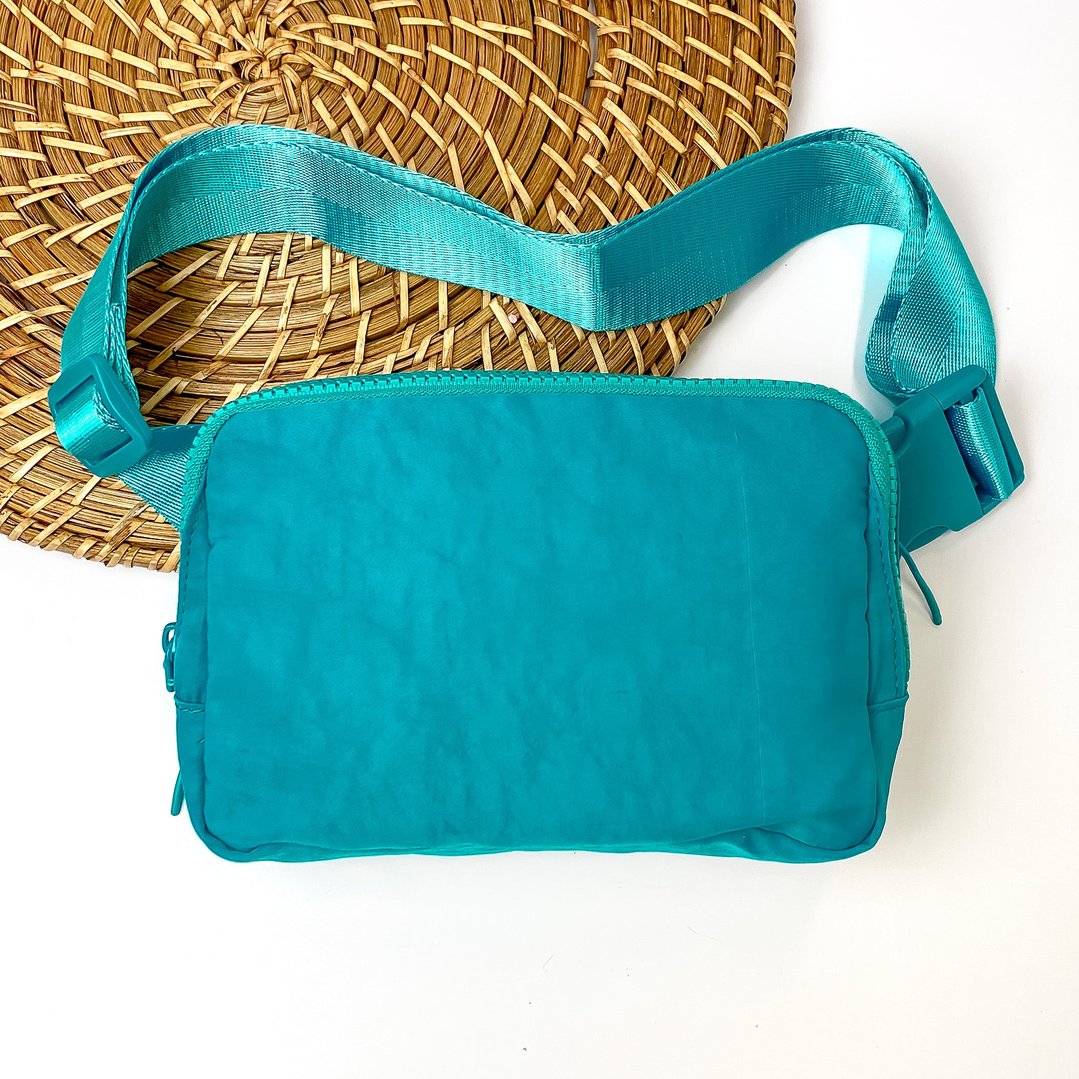 Pictured is a rectangle fanny pack with a top zipper with tassel in teal. This bag also includes a teal strap and teal accents. This bag is pictured on a white and brown patterned background. 
