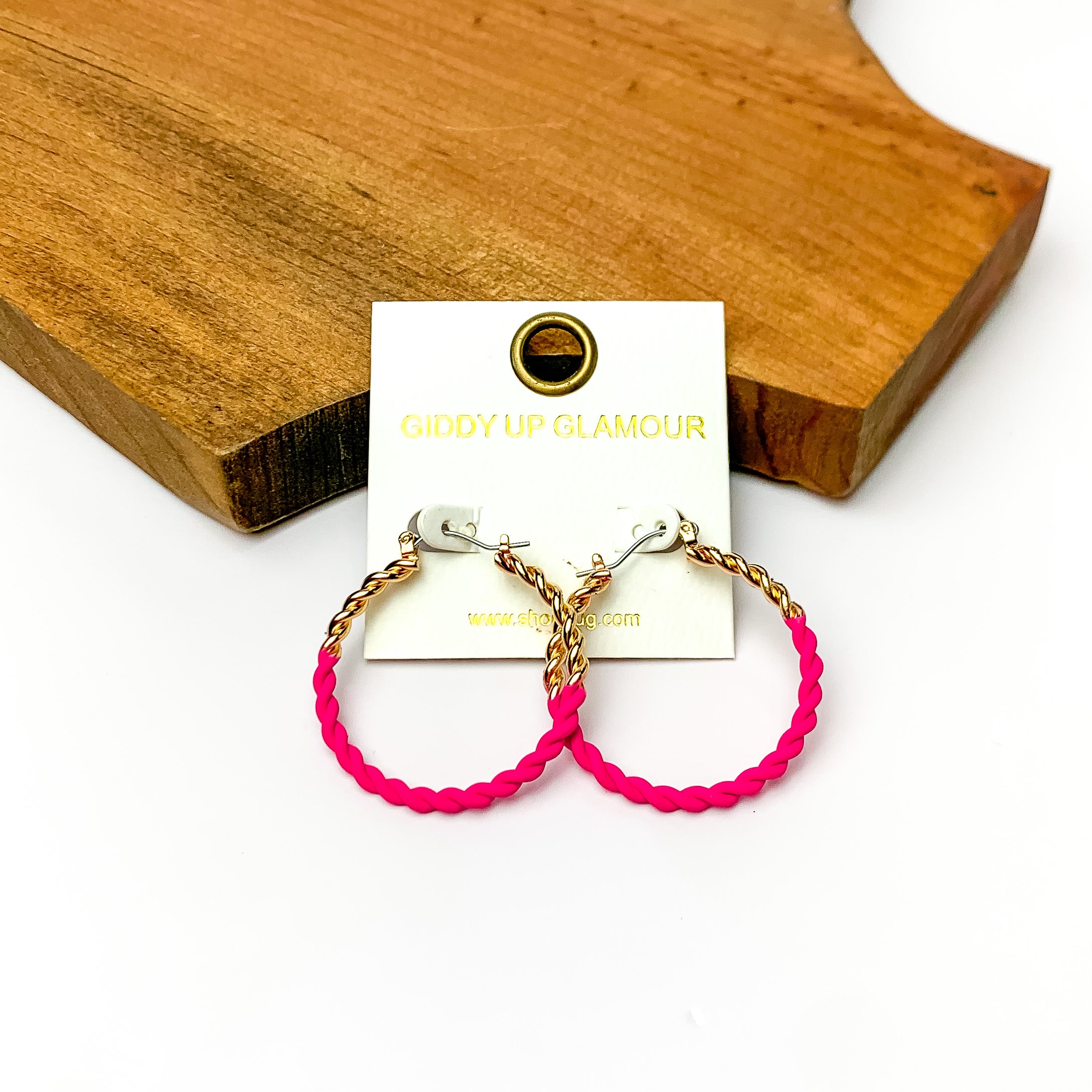 Twisted Gold Tone Hoop Earrings in Hot Pink. Pictured on a white background with wood at the top.