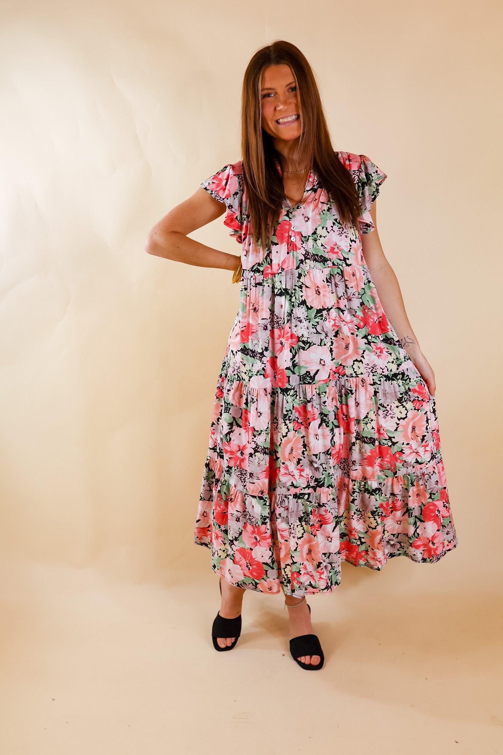 A multicolor relaxed fit midi dress. Item has a floral pattern in light pink, dark pink, pale pink with hints of dark blue/black between each flower. Also features V neckline, ruffled flowy sleeves, and pleated skirt. Item is pictured on a pale pink background