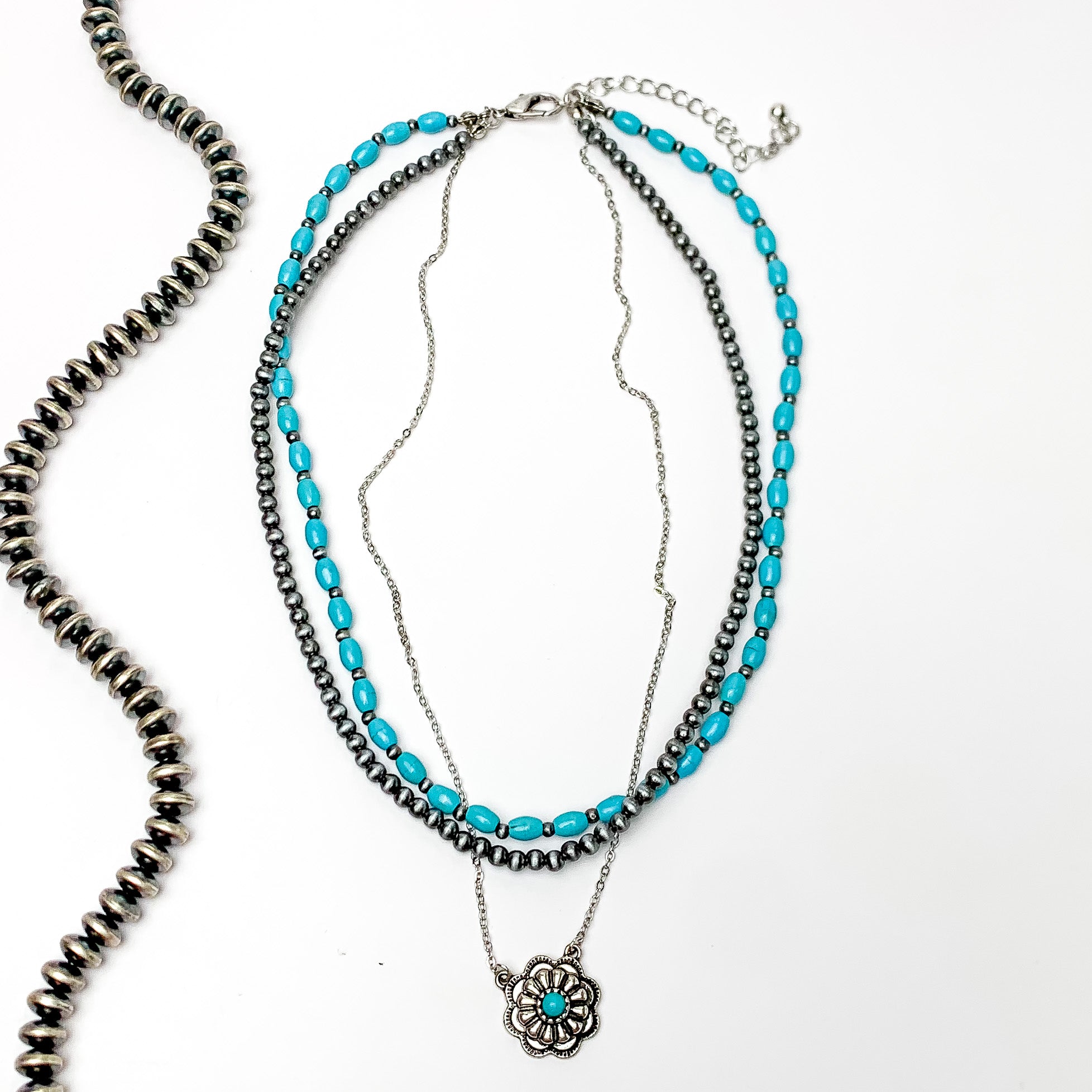 Multi strand necklace with faux navajo and turquoise beads and a dangling silver flower pendent with a turquoise stone in the middle. Pictured on a white background with a strand of silver beads to the left of it.