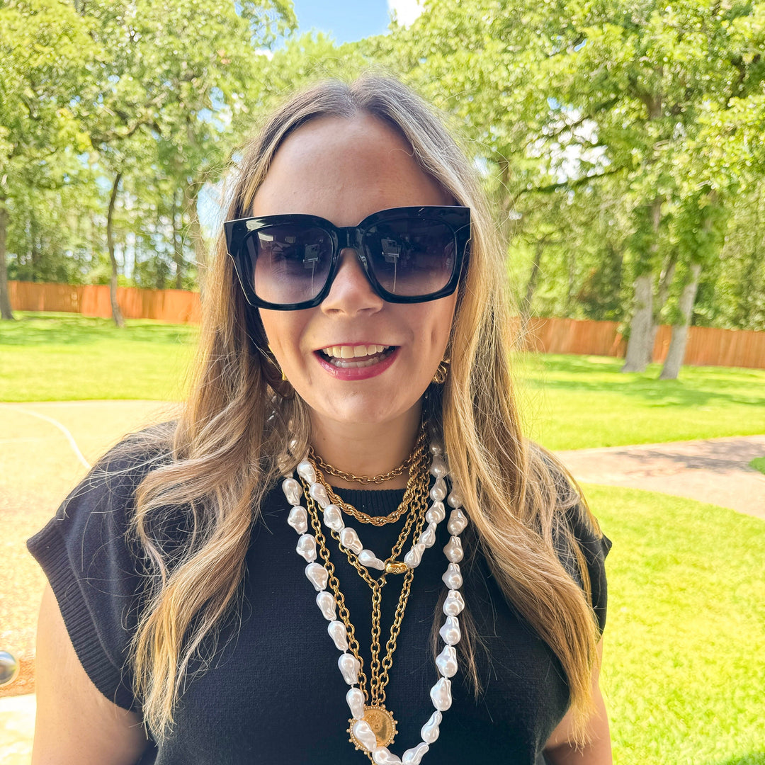 Woman wearing sunglasses and a long necklace outdoors with greenery in the background