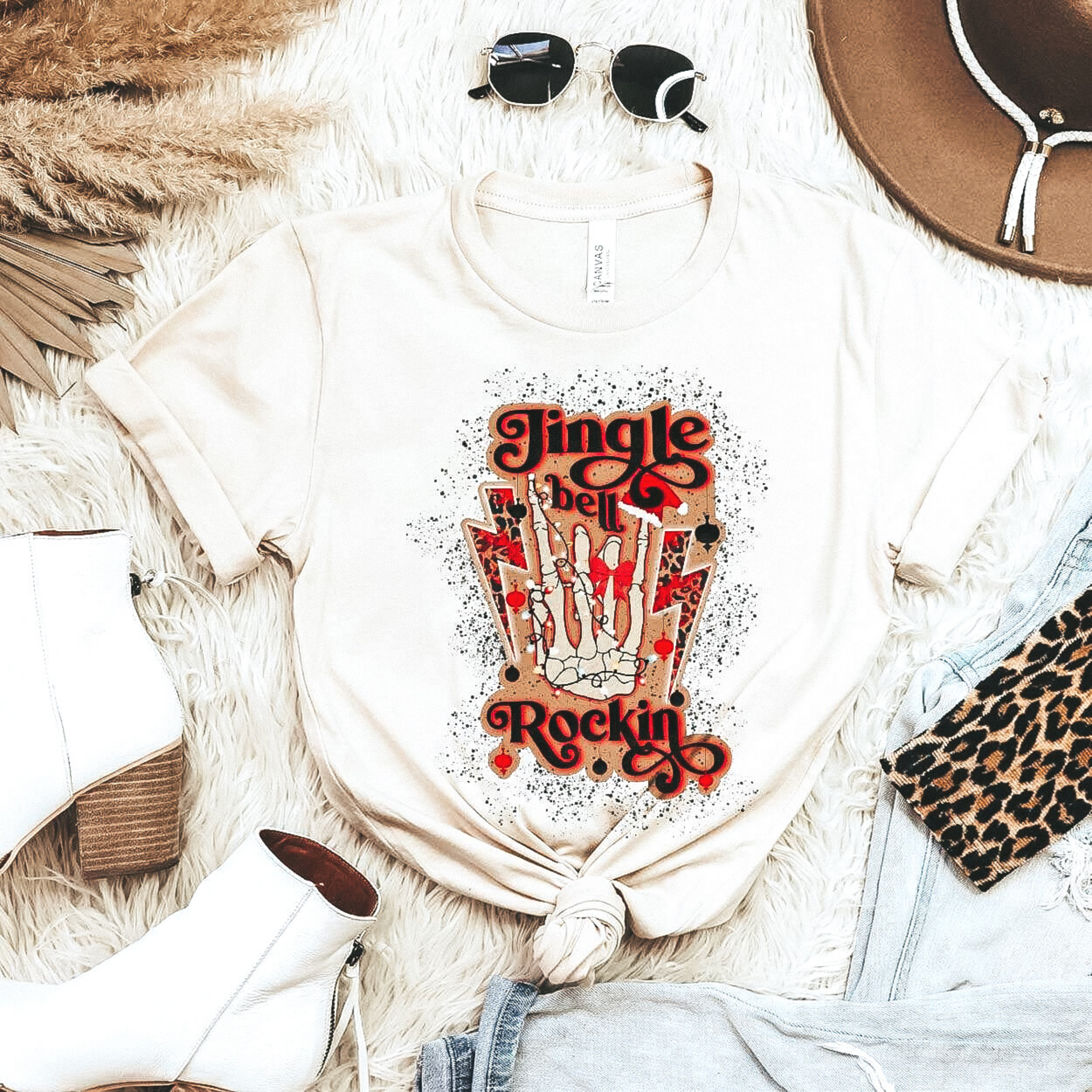 A cream colored crew neck tee with a skeleton hand making the rocker symbol that says "Jingle Bell Rockin'." Pictured with white booties, sunglasses, and a brown hat.