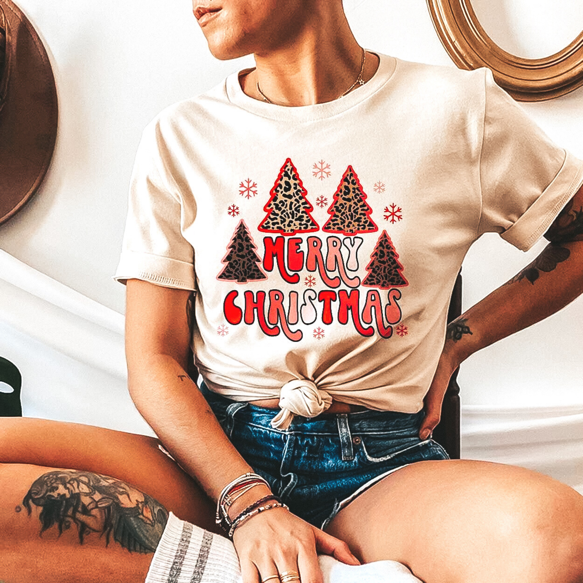 Model is sitting criss-cross wearing a short sleeve crew neck tee shirt with leopard christmas trees and the phrase "Merry Christmas" written in bubble letters. She has this tee modeled with rolled sleeves and dark wash denim shorts. 