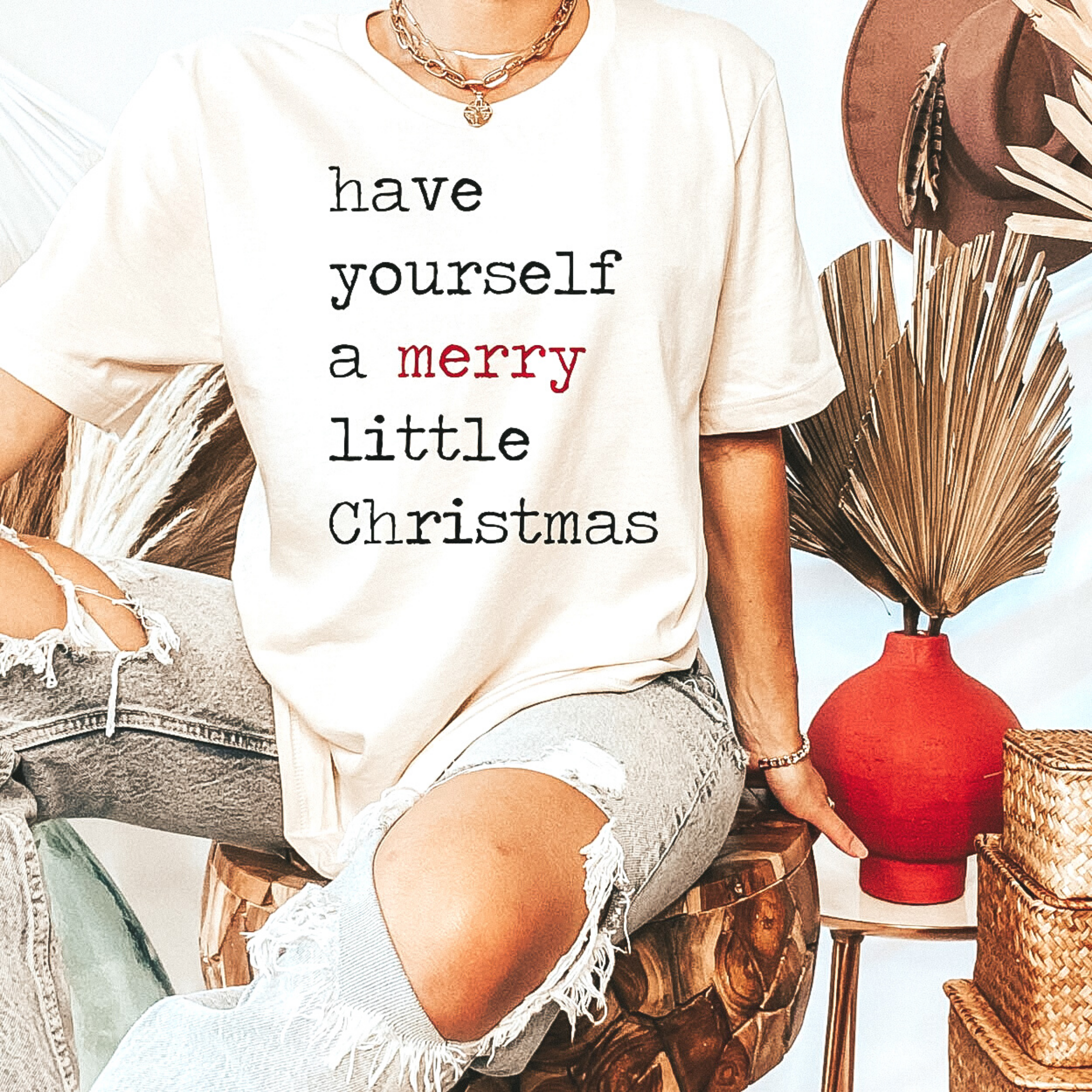 Model is wearing short sleeve, crew neck graphic tee that says "Have Yourself a Merry Little Christmas" on an ivory background. Model has it paired with distressed light wash jeans and gold jewelry.