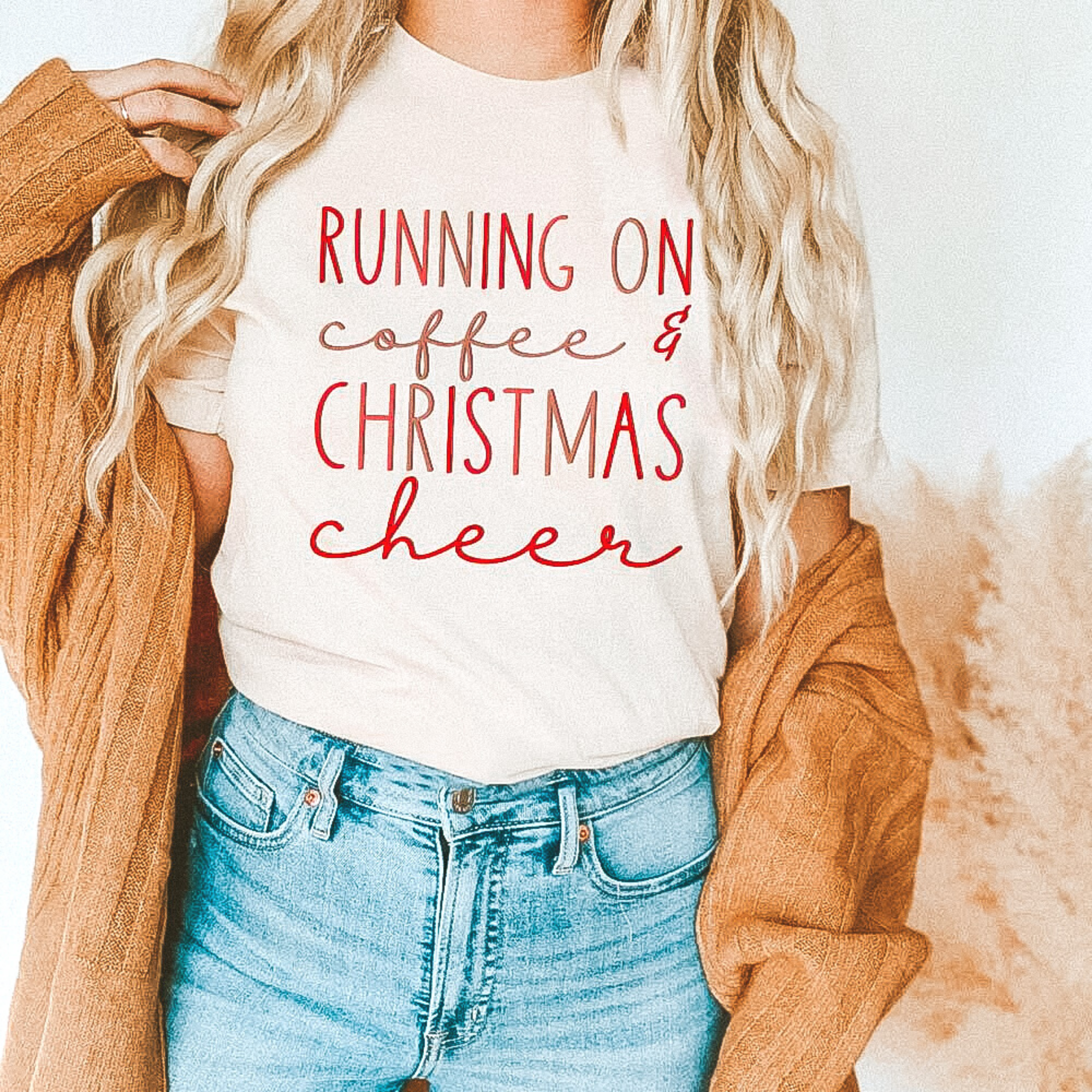 Model is wearing a short sleeve crew neck graphic tee that says "Running on Coffee & Christmas Cheer." Model has it paired with a camel brown cardigan and light wash blue jeans.