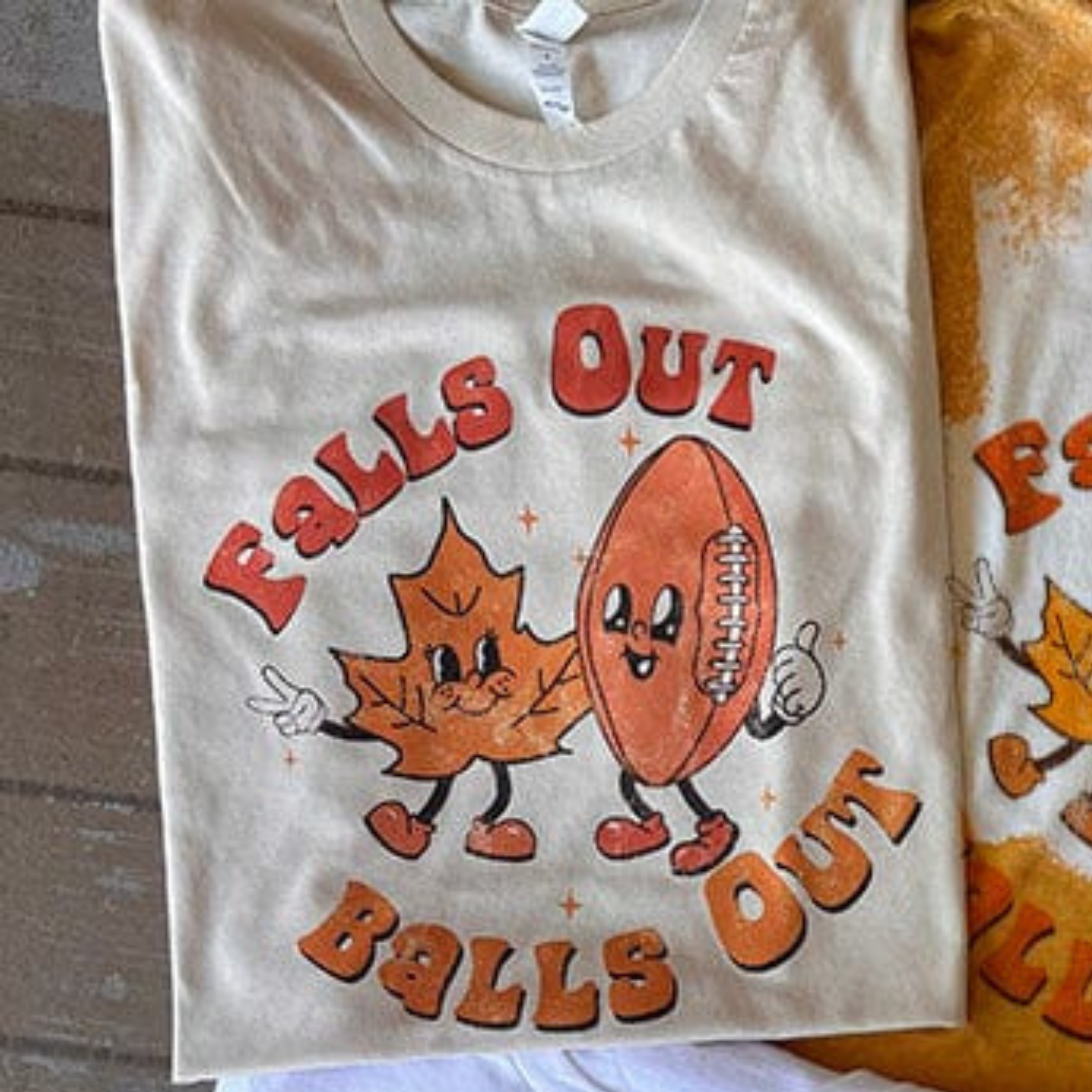 This Bella + Canvas cream tee includes a crew neckline, short sleeves, and a cute hand drawn design of an orange leaf and football with the words "Fall Out" above them and "Balls Out" below them. The fonts are in rust orange and mustard yellow, perfect for Fall. This is shown as a flat lay in this photo.