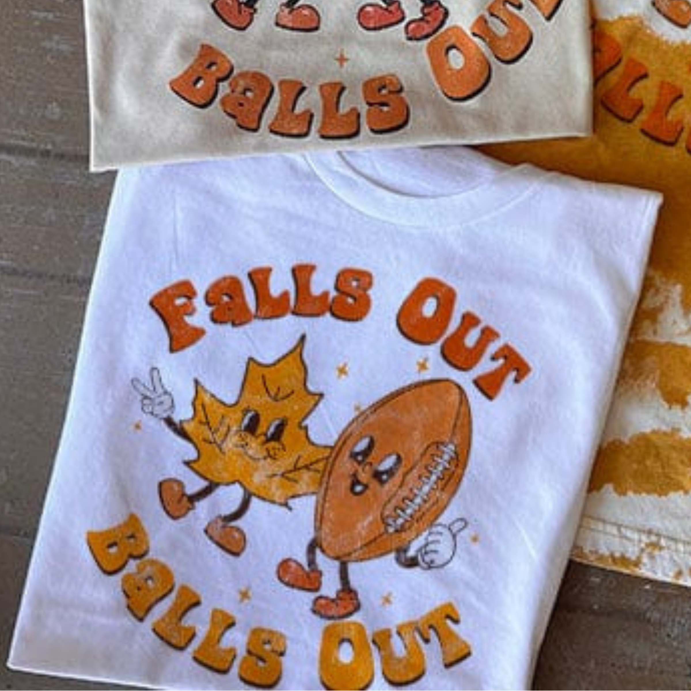 This Bella + Canvas white tee includes a crew neckline, short sleeves, and a cute hand drawn design of an orange leaf and football with the words "Fall Out" above them and "Balls Out" below them. The fonts are in rust orange and mustard yellow, perfect for Fall. This is shown as a folded flat lay in this photo.