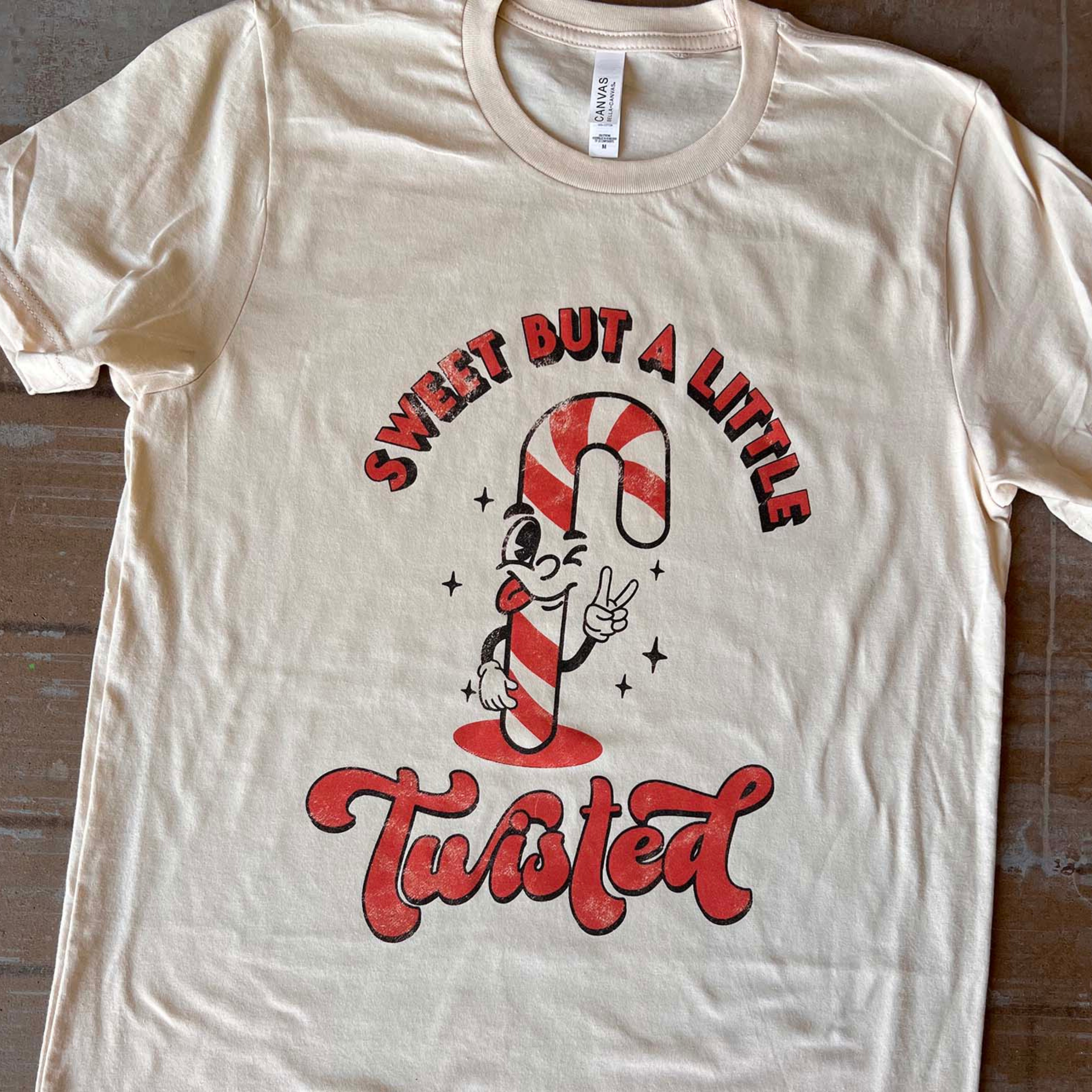 This cream Bella + Canvas tee includes a crew neckline, short sleeves, and a graphic that says "Sweet But A Little Twisted" in a mix of cute, red fonts with a candy cane throwing up the peace sign. It is pictured here as a flat lay. 