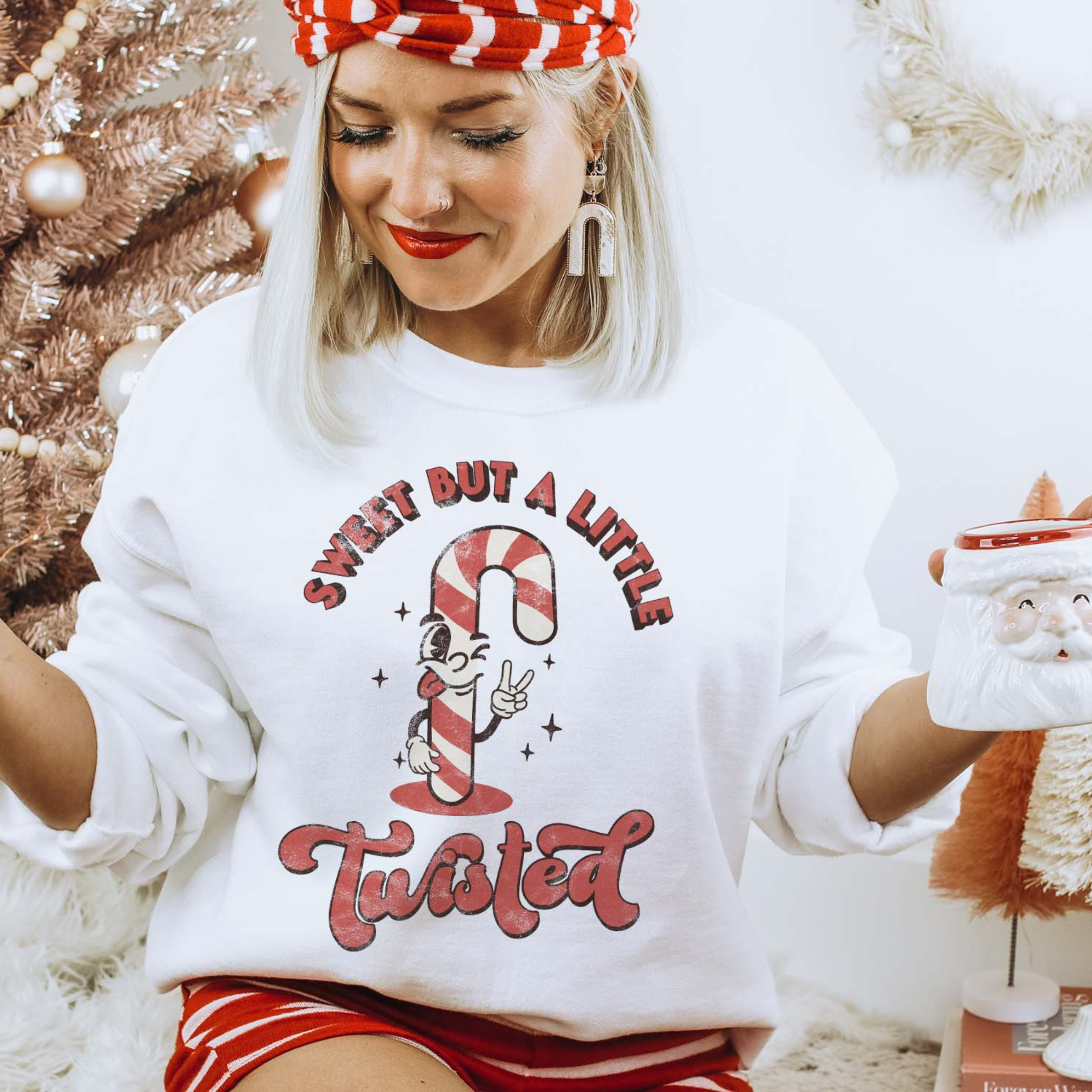 This white sweatshirt includes a crew neckline, long sleeves, and a graphic that says "Sweet But A Little Twisted" in a mix of cute, red fonts with a candy cane throwing up the peace sign. The model has this sweatshirt paired with a red and white striped headband, white earrings, and a red and white striped pair of shorts. She also has the sleeves rolled and she is holding a Santa Clause mug. 