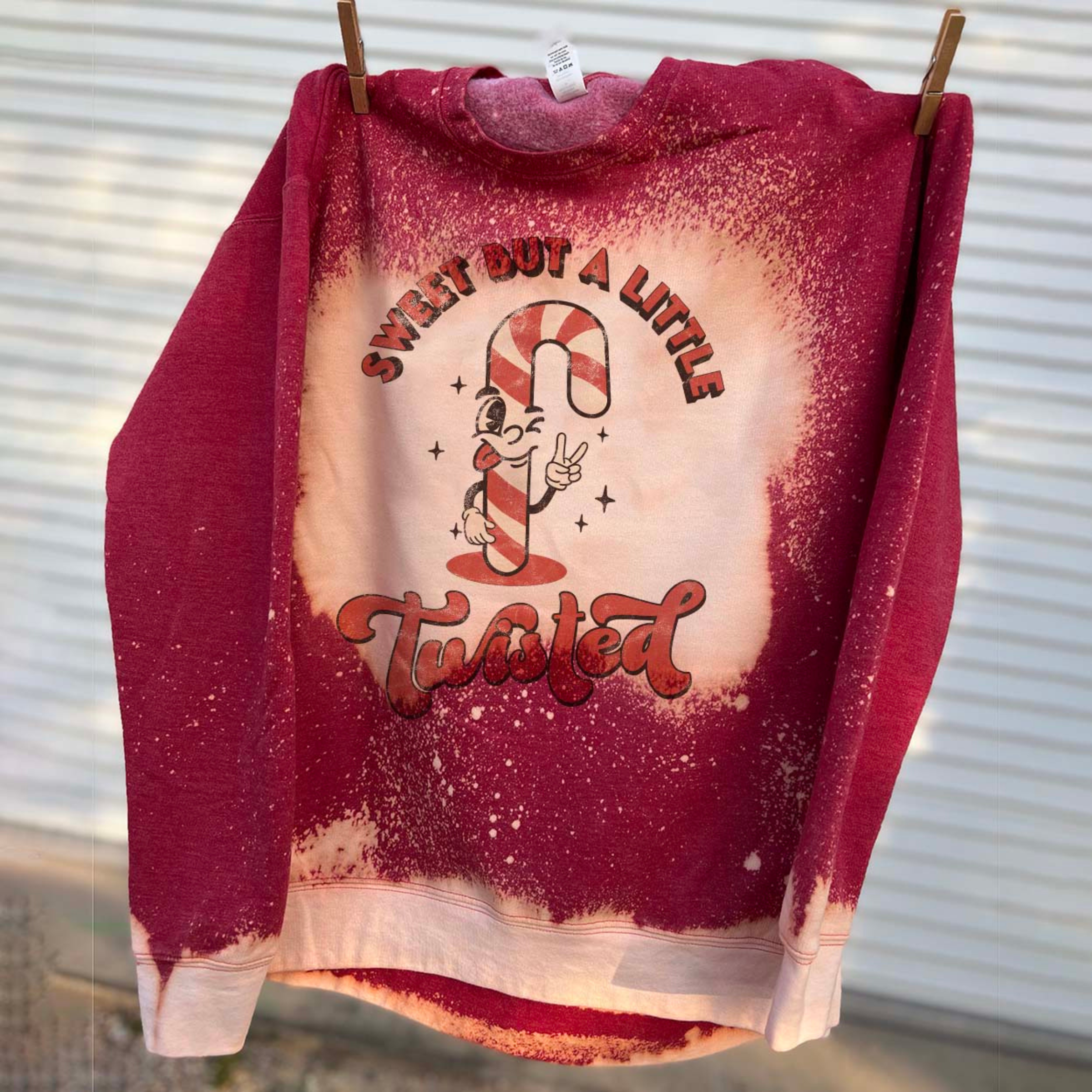 This bleached splatter graphic sweatshirt in red includes a crew neckline, long sleeves, and a graphic that says "Sweet But A Little Twisted" in a mix of cute, red fonts with a candy cane throwing up the peace sign. It is pictured here hung from a line.