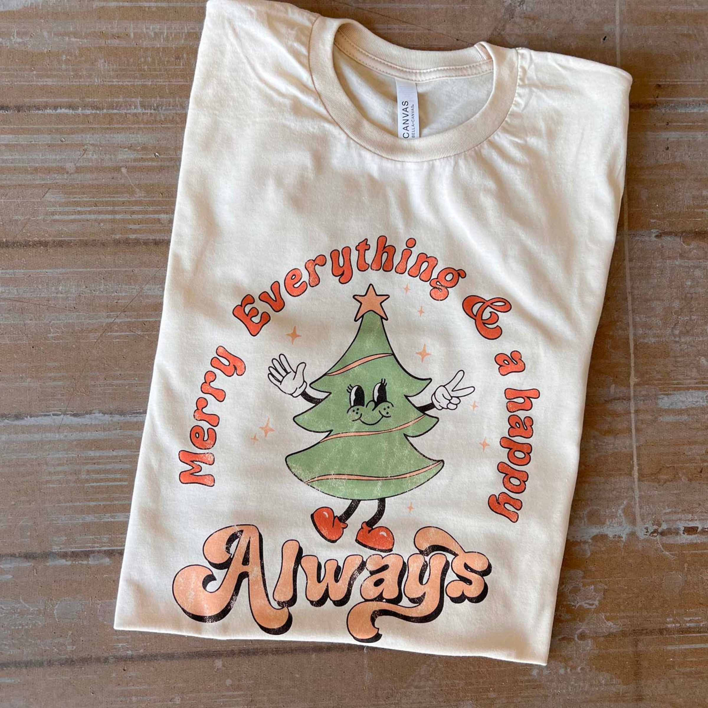 This cream Bella + Canvas tee includes a crew neckline, short sleeves, and a graphic that says "Merry Everything & a happy Always" in a cute mix of fonts with a Christmas Tree throwing up the peace sign. This tee is shown as a folded flat lay. 