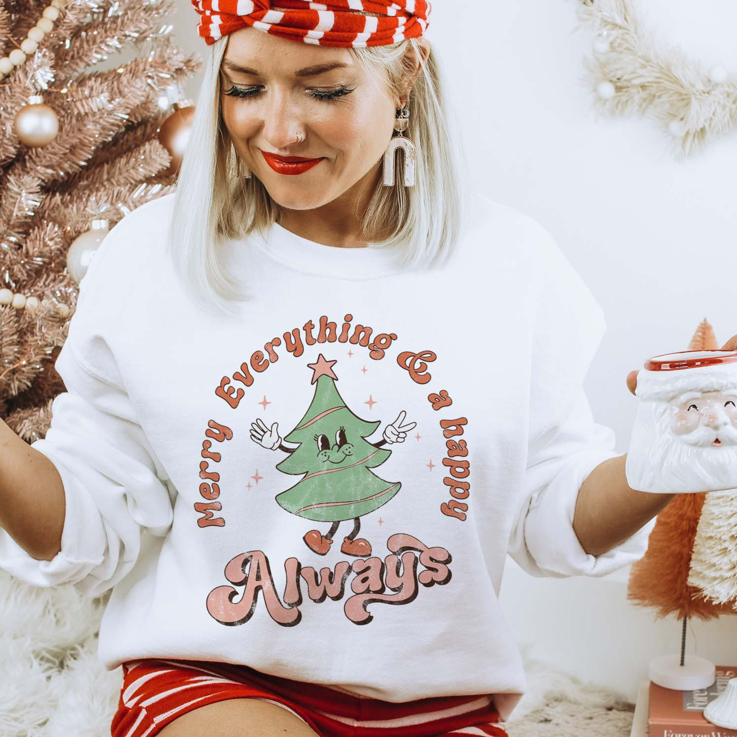 This white sweatshirt includes a crew neckline, long sleeves, and a graphic that says "Merry Everything & a happy Always" in a cute mix of fonts with a Christmas Tree throwing up the peace sign. The model has this sweatshirt paired with a red and white striped headband, white earrings, and a pair of red and white striped shorts. She also has the sleeves rolled and is holding a Santa Clause mug. 