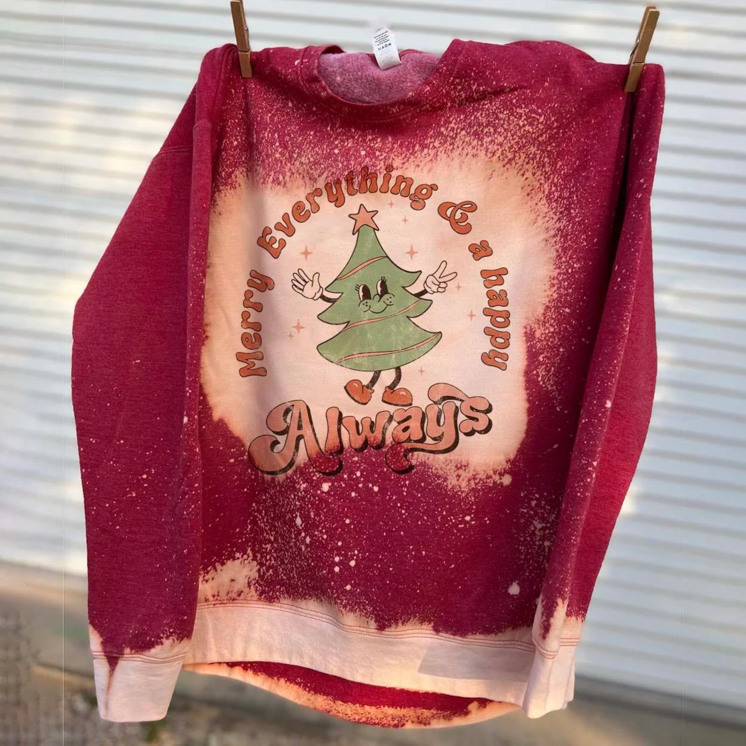 This bleached splatter graphic sweatshirt in red includes a crew neckline, long sleeves, and a graphic that says "Merry Everything & a happy Always" in a cute mix of fonts with a Christmas Tree throwing up the peace sign. This tee is shown hung from a line with clothespins. 