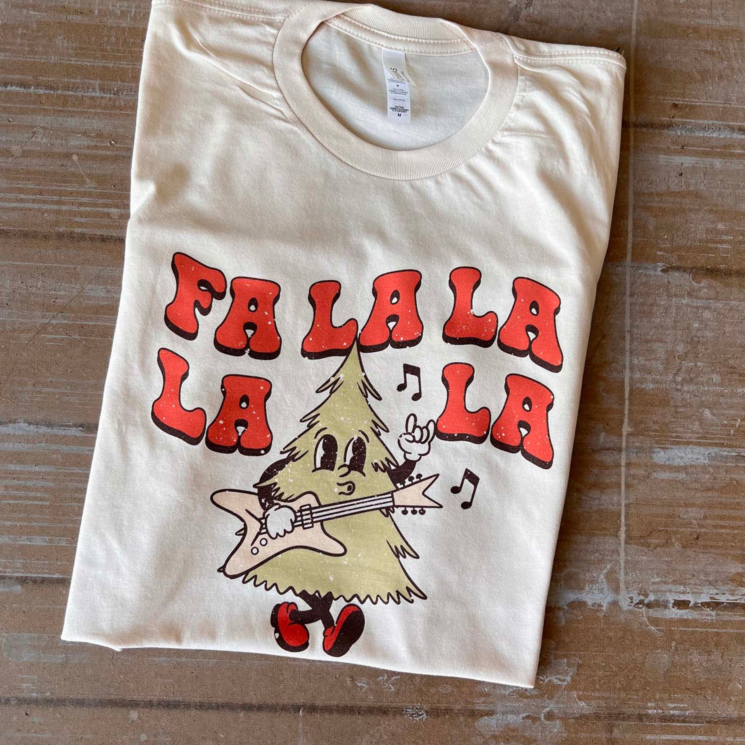 This cream tee includes a crew neckline, short sleeves, and a graphic that says "Fa La La La La" in a cute font with a Christmas Tree playing a guitar and throwing up a rock on hand symbol. This tee is shown here as a folded flat lay. 