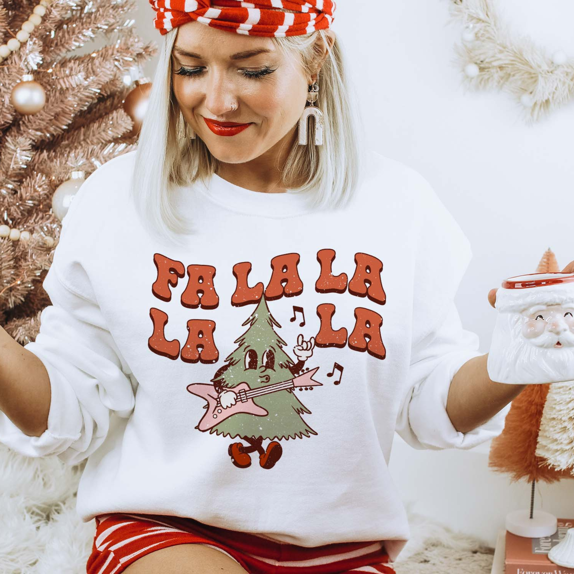 This white sweatshirt includes a crew neckline, long sleeves, and a graphic that says "Fa La La La La" in a cute font with a Christmas Tree playing a guitar and throwing up a rock on hand symbol. This sweatshirt is being modeled with a red and white striped headband, white earrings, and a pair of red and white striped shorts. The model has also rolled the sleeves of the sweatshirt and is holding a Santa Clause mug. 