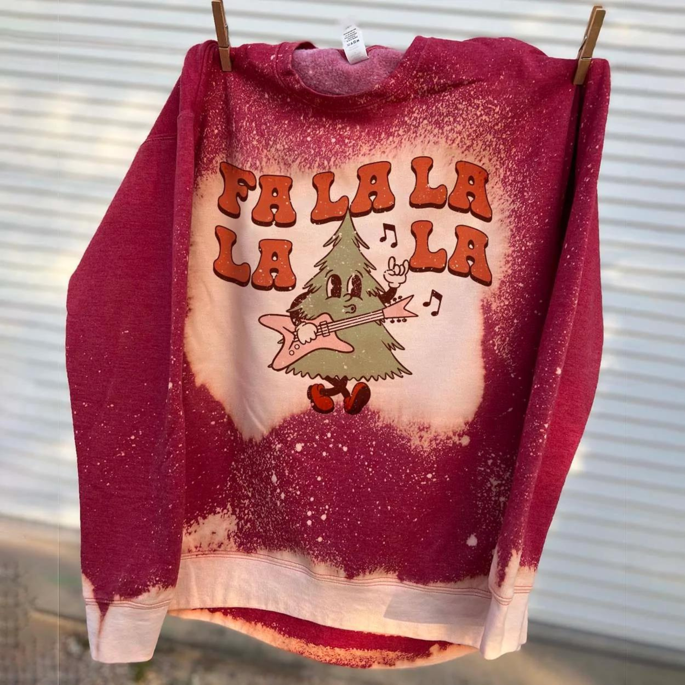 This bleached splatter graphic sweatshirt in red includes a crew neckline, long sleeves, and a graphic that says "Fa La La La La" in a cute font with a Christmas Tree playing a guitar and throwing up a rock on hand symbol. This sweatshirt is shown here hanging on a clothing line with clothespins. 