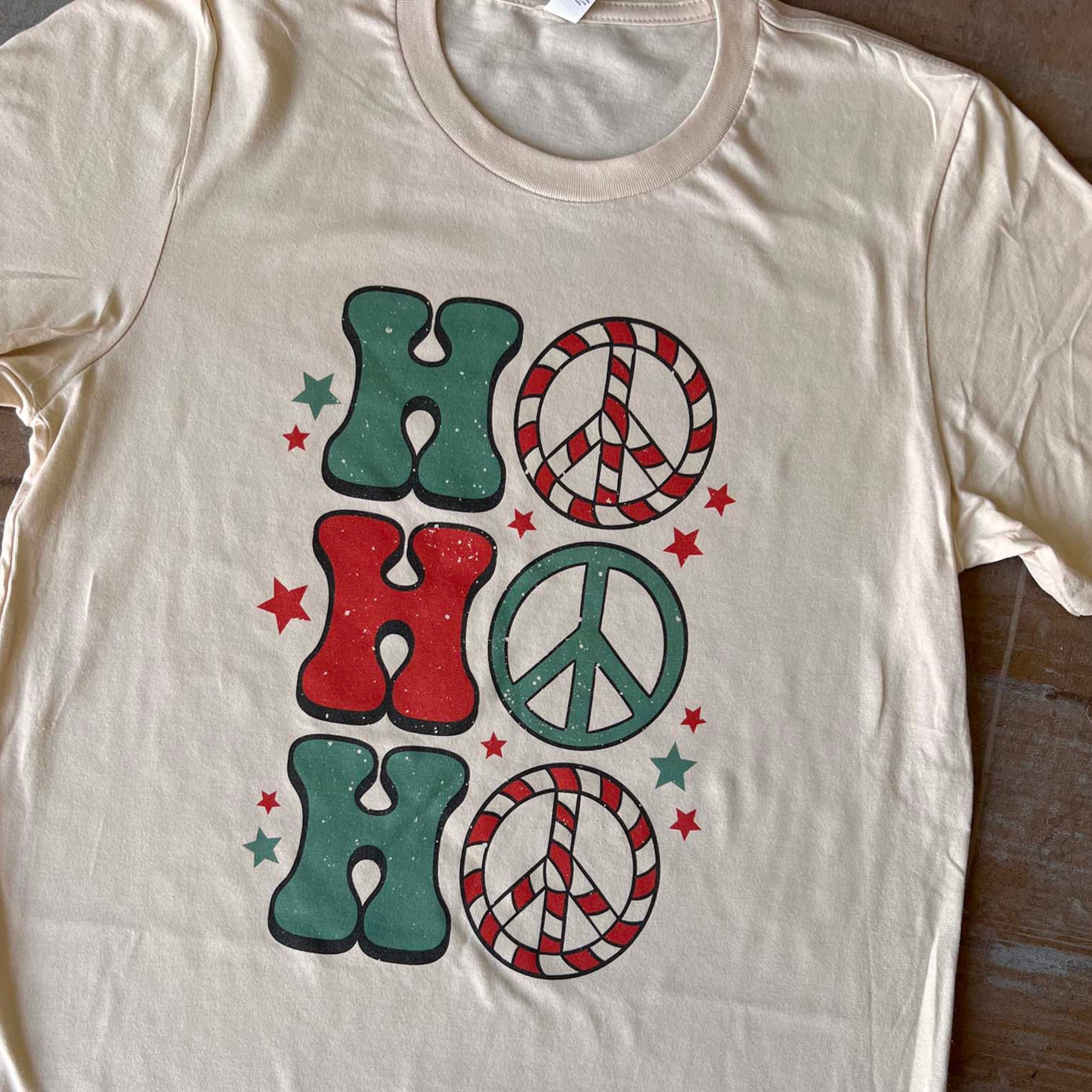 This cream tee includes a crew neckline, short sleeves, and a graphic that says "Ho Ho Ho" in a mix of cute font in red and green. The O's are peace signs and stars are all around the graphic. This tee is shown here as a flat lay. 