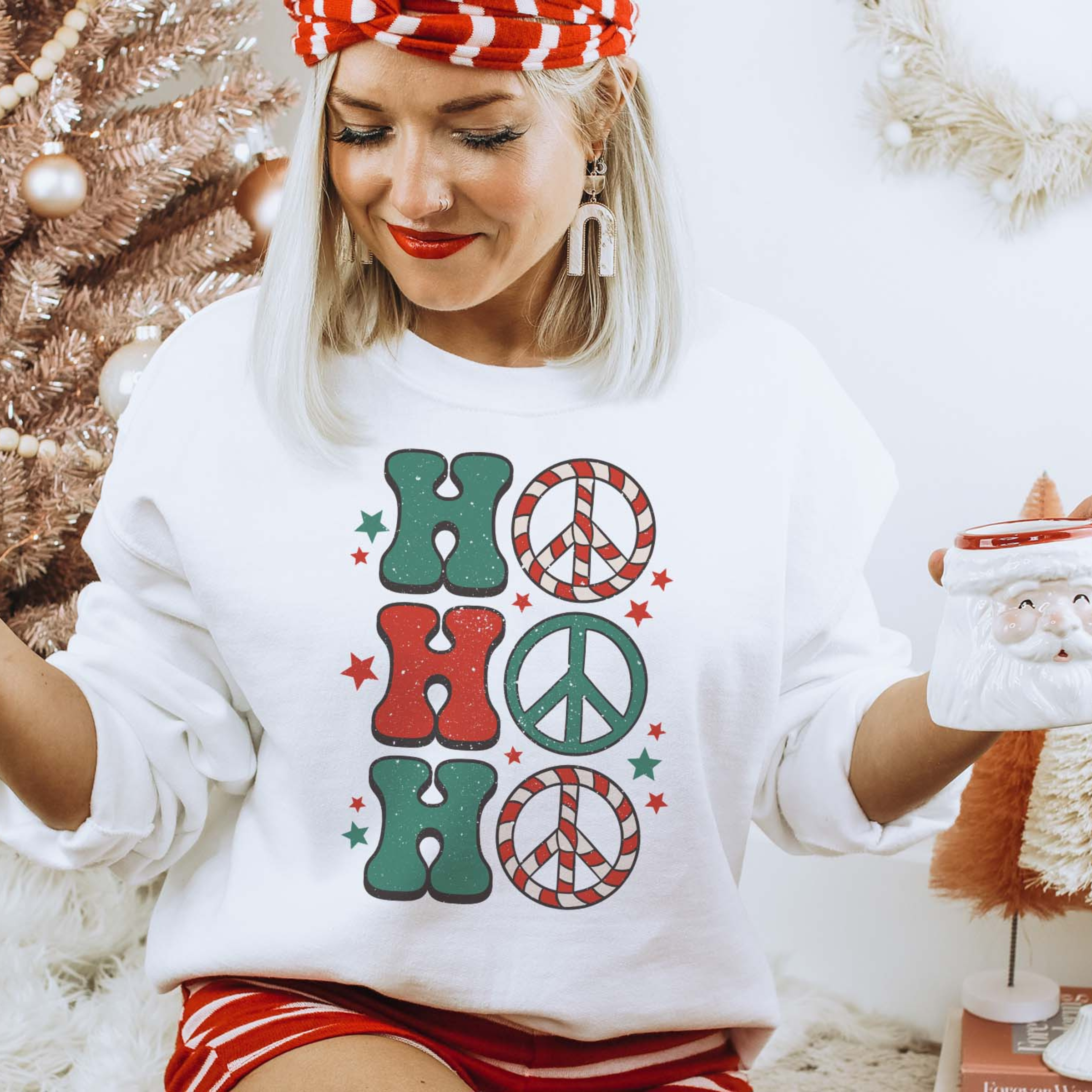 This white sweatshirt includes a crew neckline, long sleeves, and a graphic that says "Ho Ho Ho" in a mix of cute font in red and green. The O's are peace signs and stars are all around the graphic. This is modeled with a red and white striped headband, white earrings, and a pair of red and white striped shorts. The model has rolled the sleeves and is also holding a Santa Clause mug. 