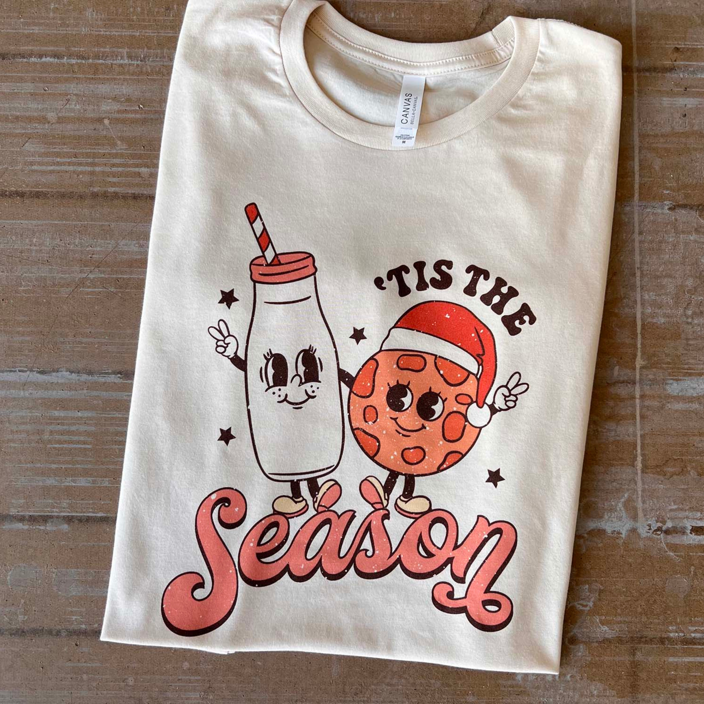 This cream tee includes a crew neckline, short sleeves, and a graphic that says " 'Tis the Season" in a cute font with a cartoon milk and cookie wearing a Santa hat; both are throwing up peace signs with their outer hands and looking at each other. This tee is shown here as a flat lay. 