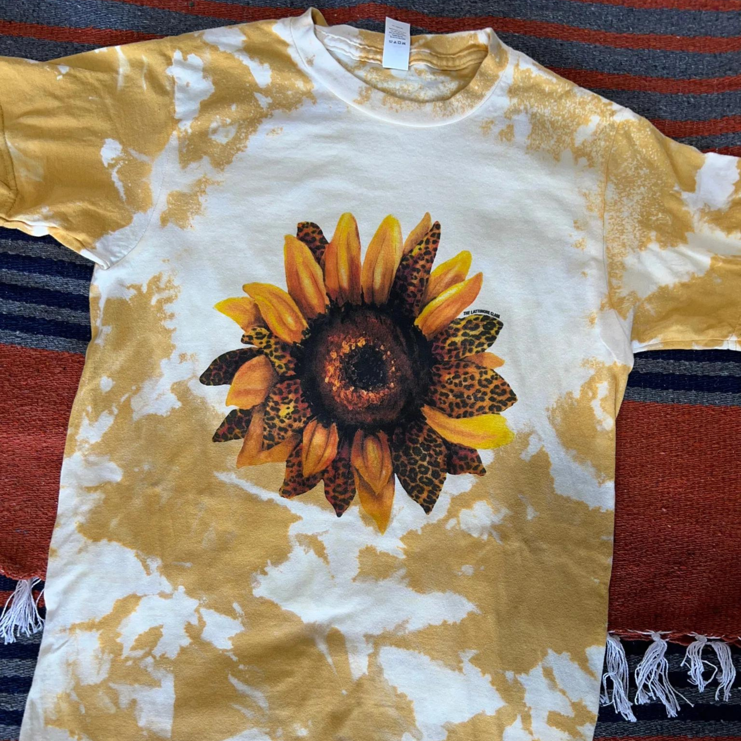 This mustard yellow bleached tee includes a crew neckline, short sleeves, and a cute graphic of a leopard sunflower. It is pictured as a flatlay on a red. gray, and black striped serape blanket background. 