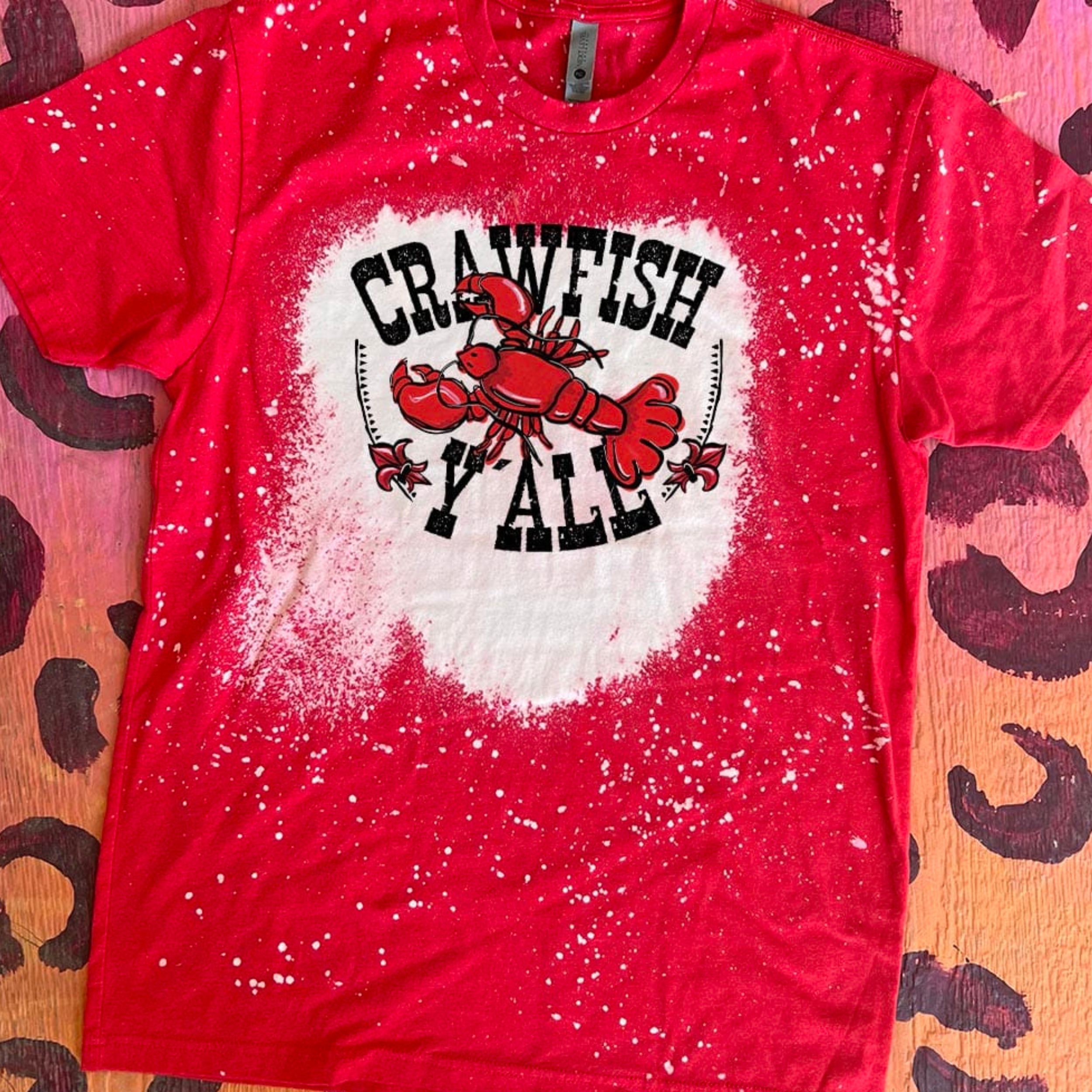 A red tee shirt with short sleeves and a crew neckline with a bleach splatter graphic crawfish that says "crawfish y'all." This tee shirt is pictured on a leopard print background.