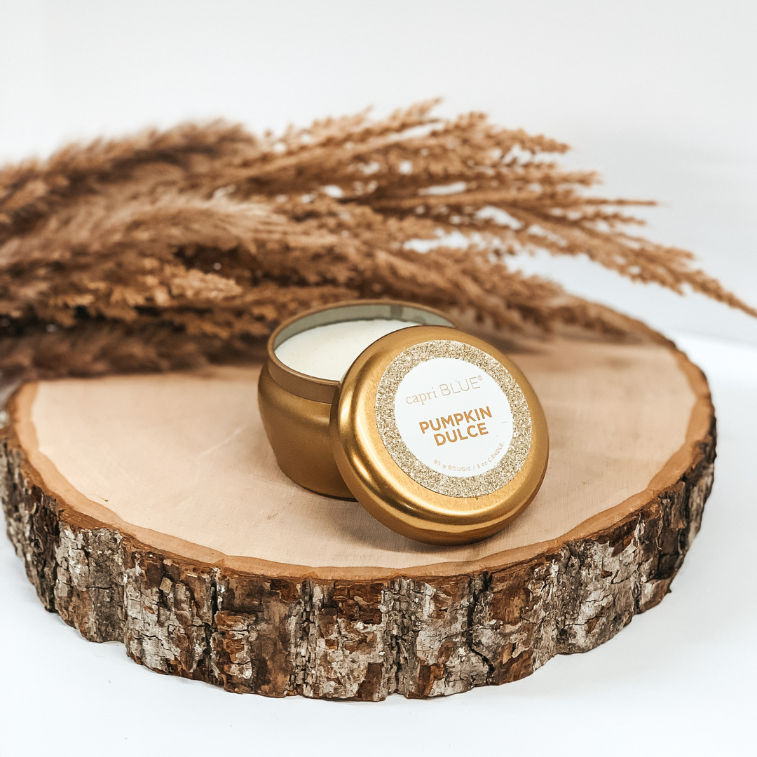 Gold tin candle with a "Caprie Blue Pumpkin Dulce" sticker on the lid pictured on a piece of wood on a white background. 