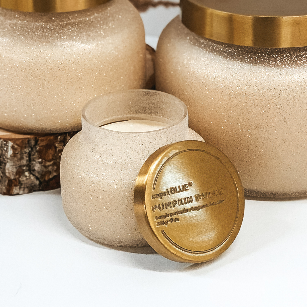 Champagne glitter candle with a gold lid pictured on a white background in front of two champagne glitter candles. 