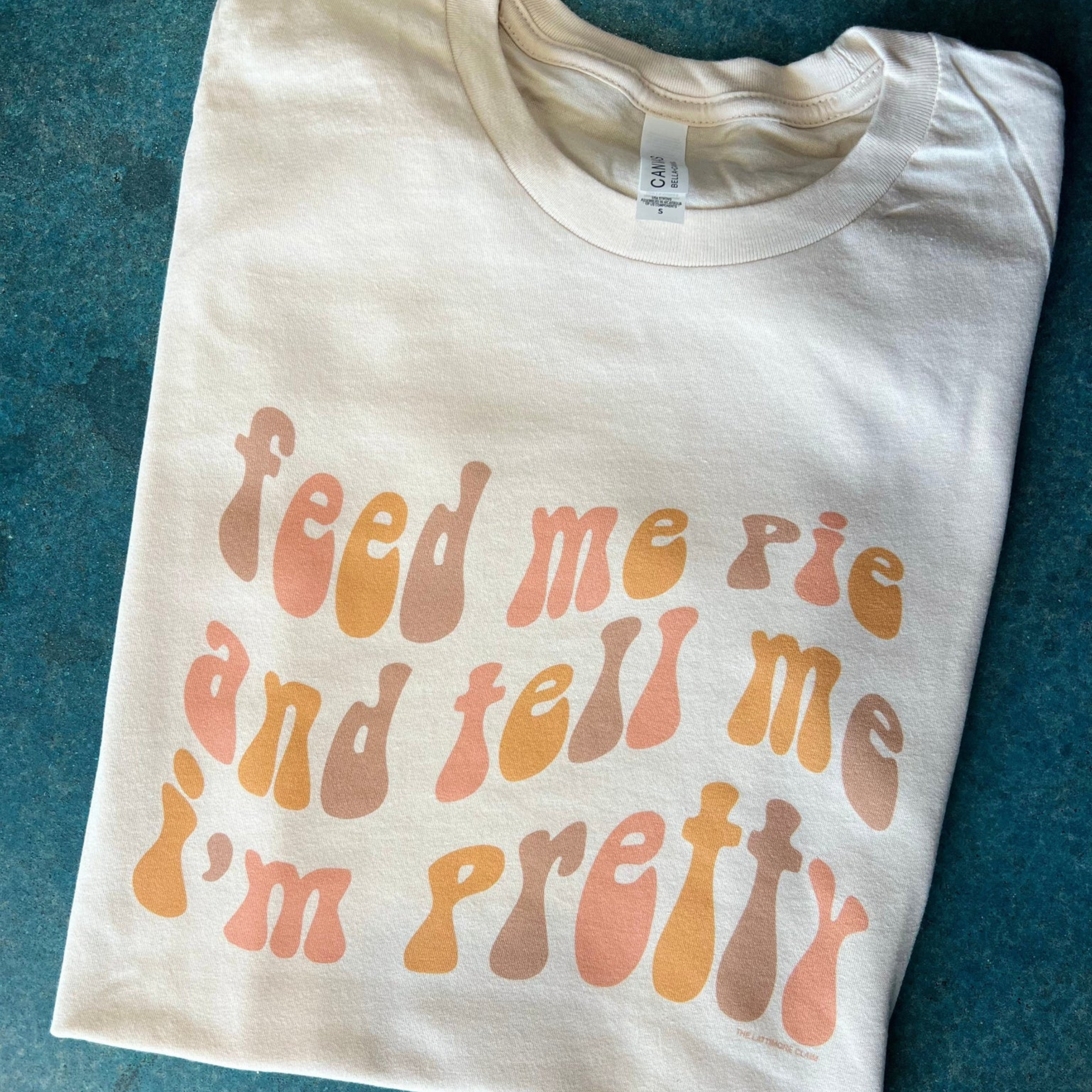 Cream tee folded in half on blue background with groovy words saying "feed me pie and tell me i'm pretty" in colors brown, pink and mustard alternating.