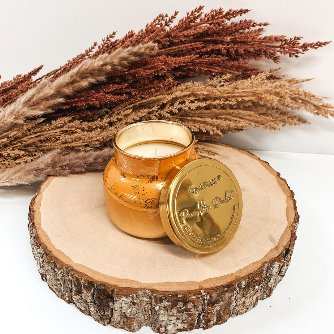 Orange candle with gold flecks and a gold lid pictured on a white background on a piece of wood with brown pompous grass in behind the candle. 