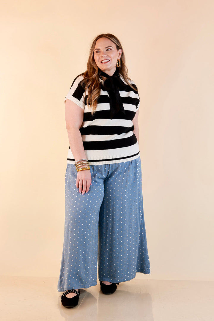 Model wearing an ivory and black striped short sleeve sweater polo with a sheer black scarf tied at the neck, light blue polka dot wide-leg pants and black flats.
