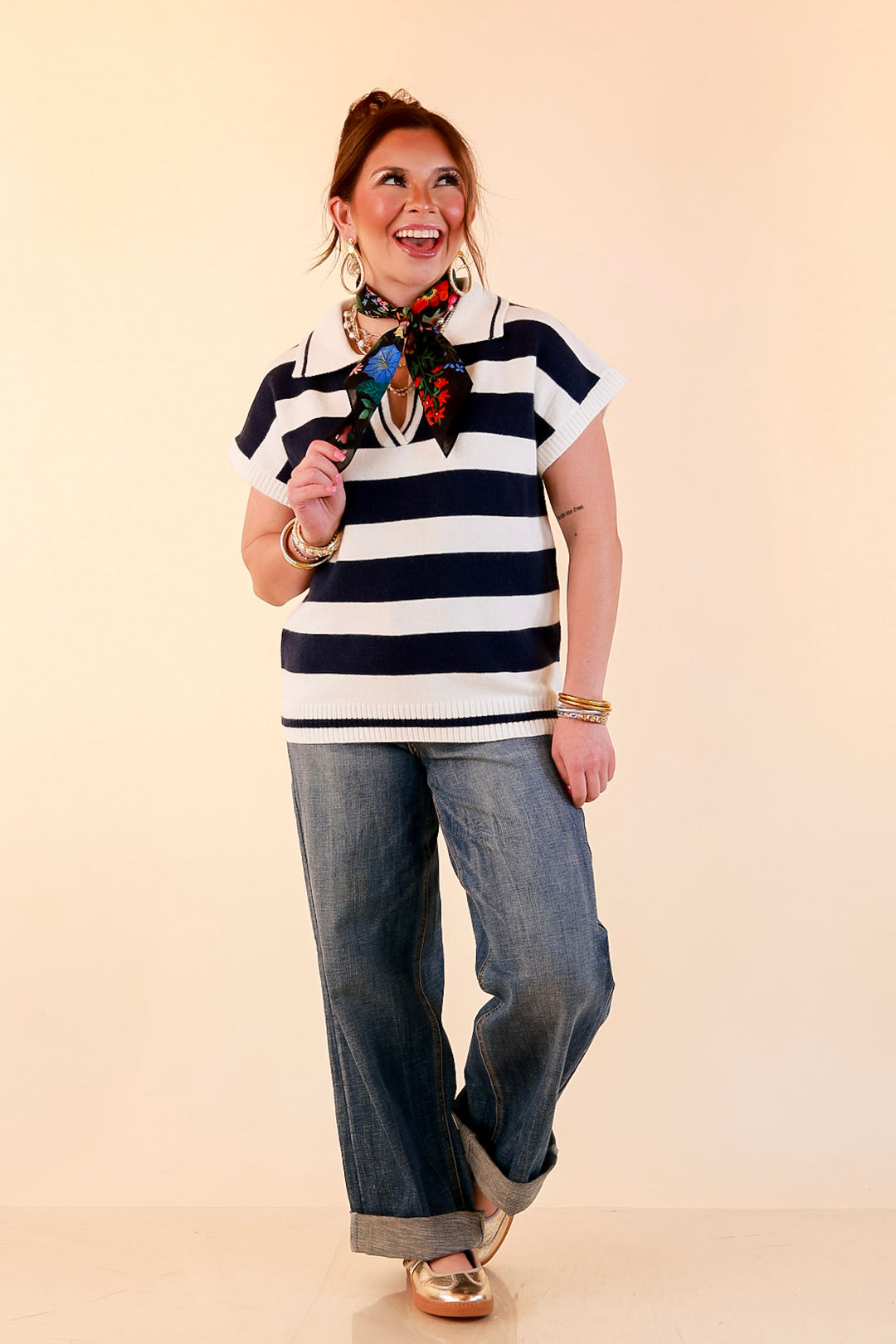 Model wearing an ivory and navy striped short sleeve sweater polo with a black floral scarf tied at the collar, cuffed medium wash jeans and gold flats.