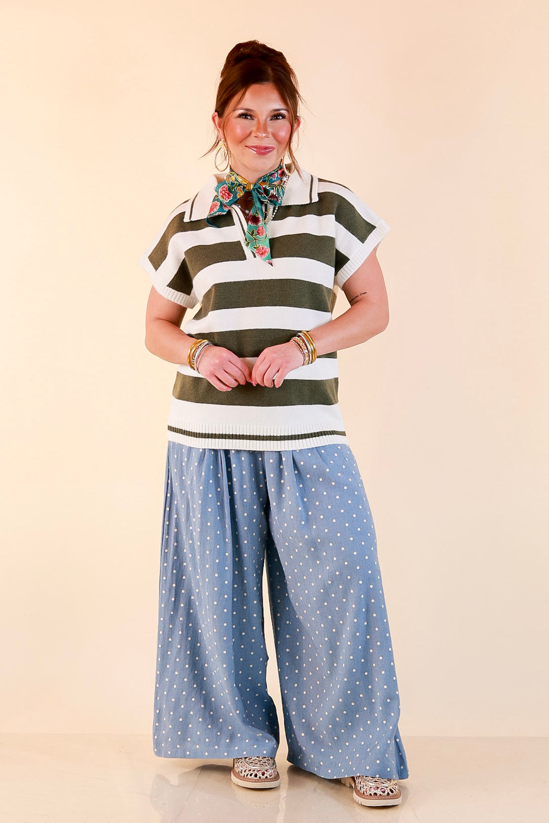 Model wearing an ivory and olive green striped short sleeve sweater polo with a printed scarf tied at the collar and light blue polka dot wide-leg pants.
