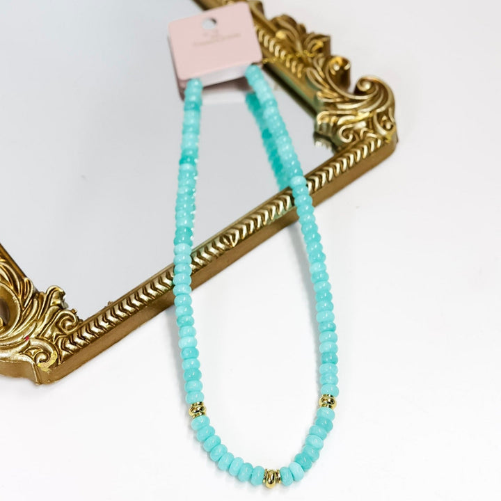 Turquoise beaded necklace with gold accents on a white surface with a decorative gold frame.