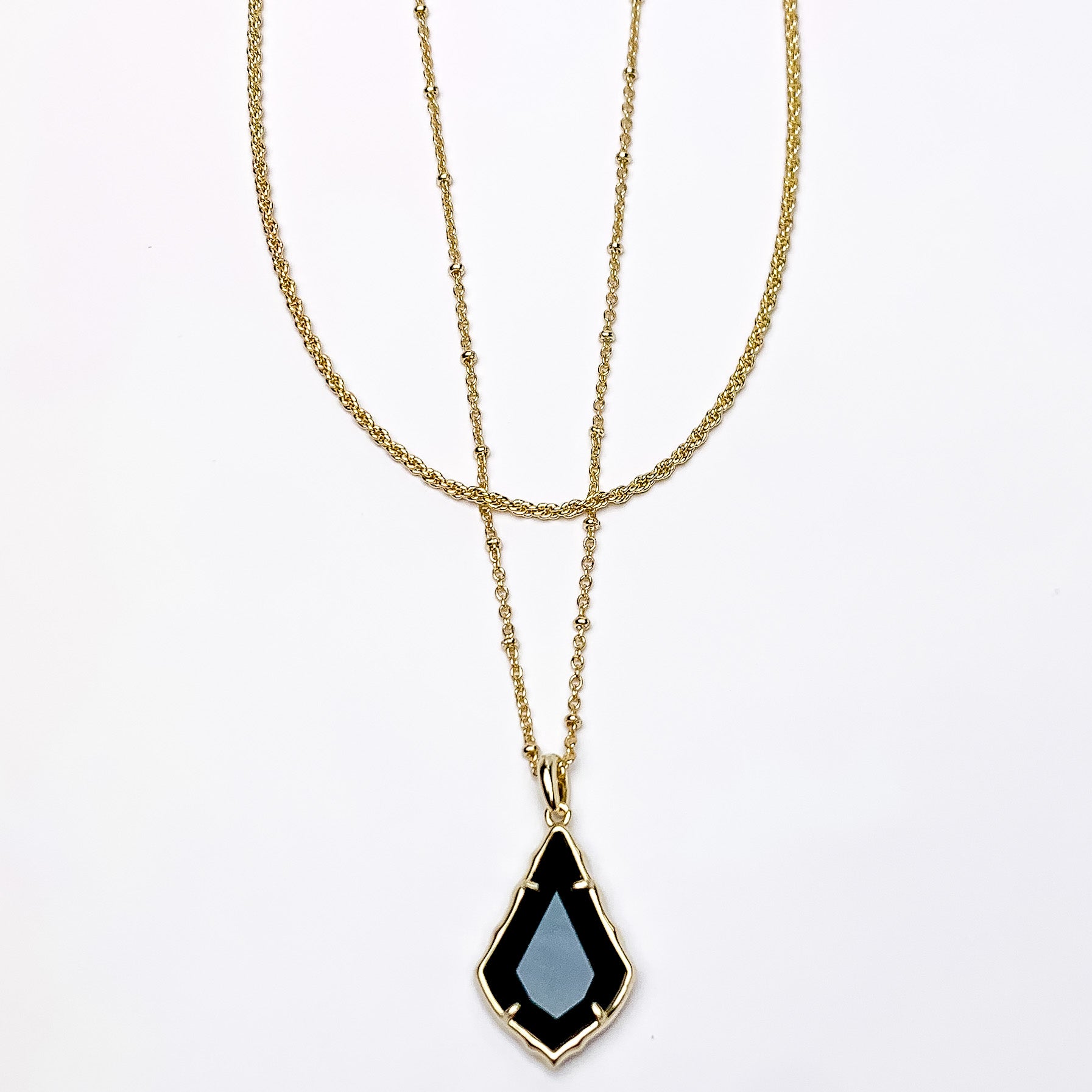 Two gold chain necklaces with the longer strand that has a gold and black opaque glass pendant. This necklace is pictured on a white background. 