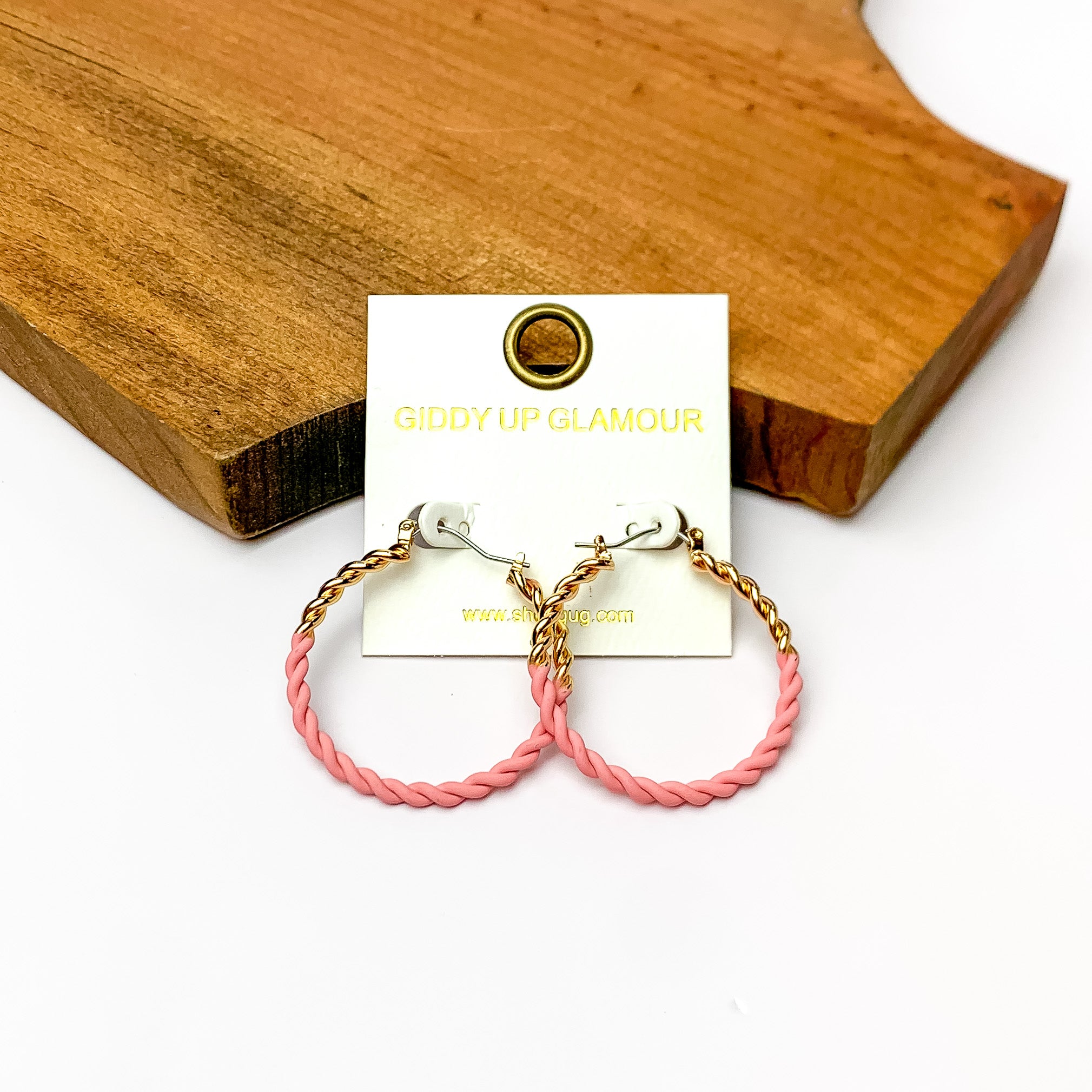 Twisted Gold Tone Hoop Earrings in Light Pink. Pictured on a white background with a wood piece behind the earrings.