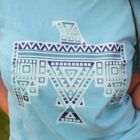 Light blue t-shirt with a thunderbird geometric pattern worn by a person outdoors.