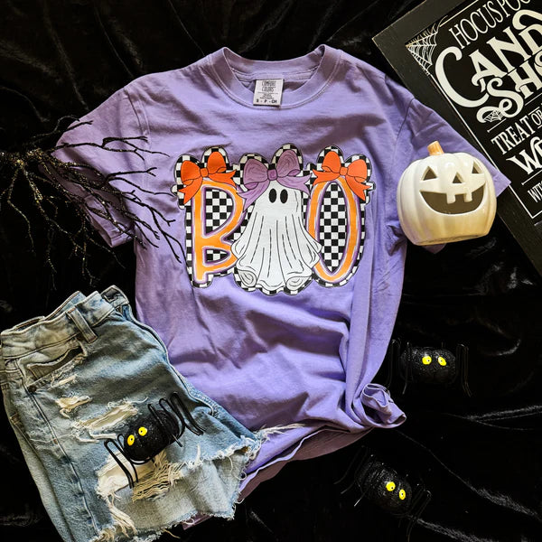 Purple t-shirt with Halloween-themed design featuring ghosts and 'BOO' text, paired with denim shorts and a small pumpkin on a dark background.