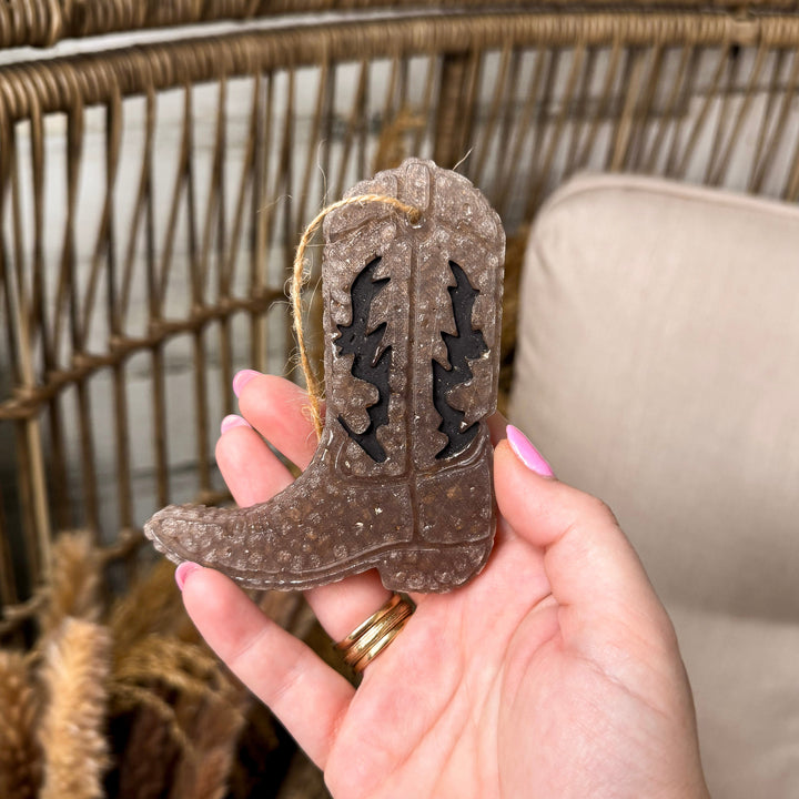 A rustic background with a hand holding a wax cowboy boot freshie.