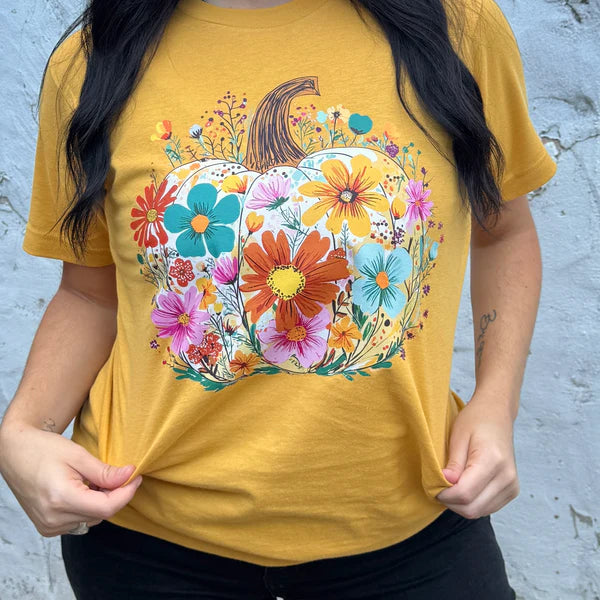 Person wearing a mustard yellow t-shirt with a floral pumpkin design against a light blue wall.